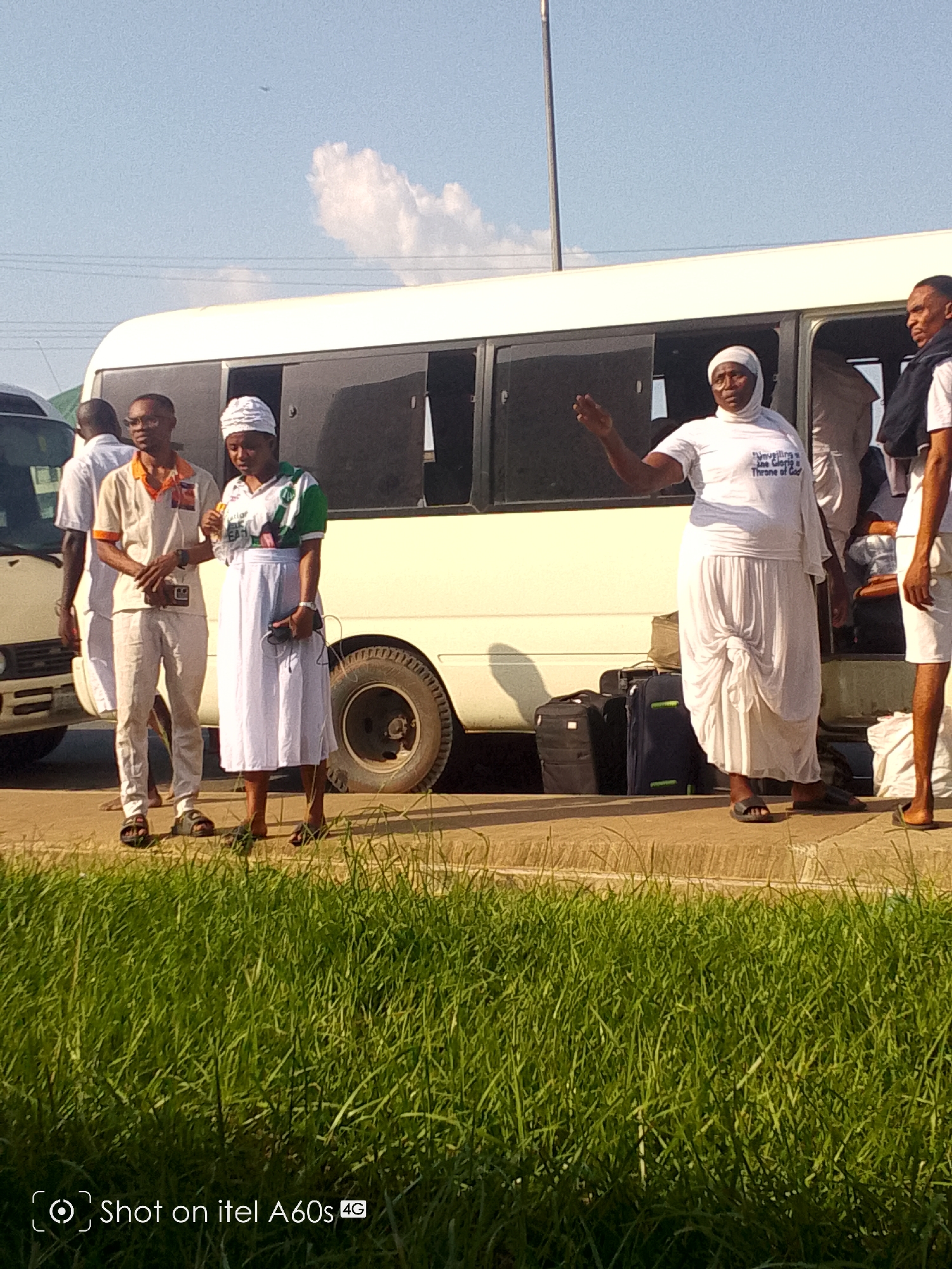 The image depicts a daytime scene in what appears to be Ikot Ekpene, Nigeria, showing a group of people gathered around a white minibus or van. The sky is bright blue with scattered white clouds, indicating clear weather. Lush green grass occupies the foreground, separated by a paved surface where the people are standing.
Several adults are the primary subjects. On the left, a man in a tan and orange polo shirt, light trousers, and glasses is visible, holding a smartphone. Next to him, a woman wearing a white head covering, a green and white patterned shirt, and a white skirt also holds a phone. Towards the right, another woman, similarly adorned with a white head covering, a white t-shirt, and a long, layered white skirt with a distinctive tied front, is actively gesturing with her right arm raised, seemingly speaking or directing. Her t-shirt bears partial text that reads, "...Unveiling the True Glory & Throne of God."
Behind this woman, dark suitcases are stacked on the ground beside the open side door of the white minibus, suggesting that the group is either preparing for a journey or has recently arrived. To the far right, a man in a white shirt and shorts stands near the bus, with a dark blue garment draped over his shoulder. Other individuals are partially visible within or near the vehicle.
The activity suggests a travel event, possibly a group outing or a religious gathering given the text on one woman's shirt. The bright sunlight and the visible shadows indicate it is daytime. The image includes a watermark in the bottom left corner, stating "Shot on itel A60s 4G".
