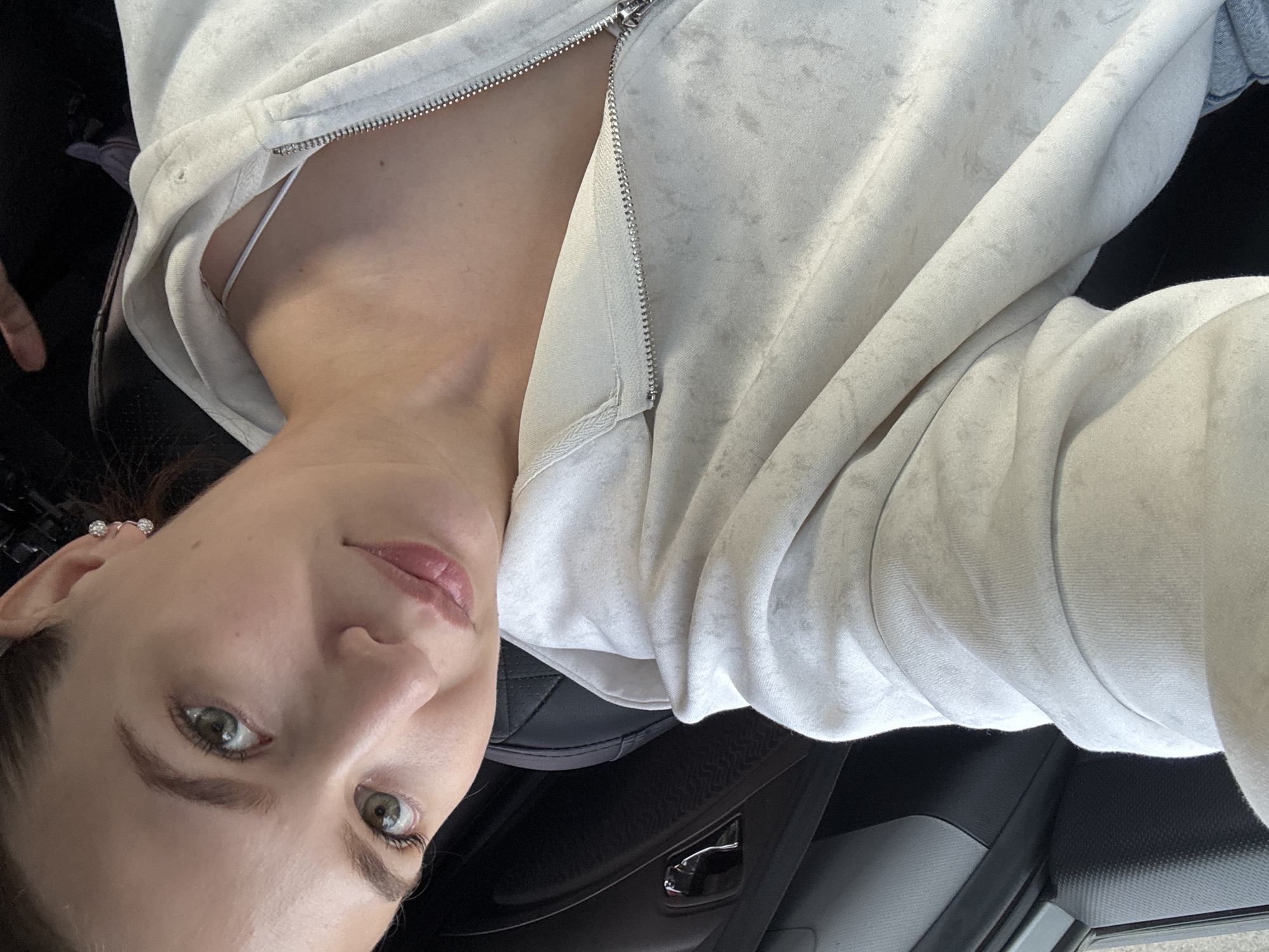 The media file is a close-up, portrait-style selfie taken from a slightly high angle, with the image rotated 90 degrees clockwise. The subject is a young woman with fair skin, green eyes, and pink lips. She is wearing a light-colored, possibly cream or off-white, zippered hoodie or jacket. A hint of a white undergarment is visible at the neckline. She has on small, sparkling earrings in her left ear. The background suggests an indoor setting, possibly a car, with dark-colored seats and interior paneling visible. The lighting appears natural and is casting soft shadows, indicating it could be daytime. The woman's expression is neutral, and her gaze is directed towards the camera. No text is visible in the image. The provided location context of Chaikovsky, Russia, does not offer specific visual cues within the image itself.