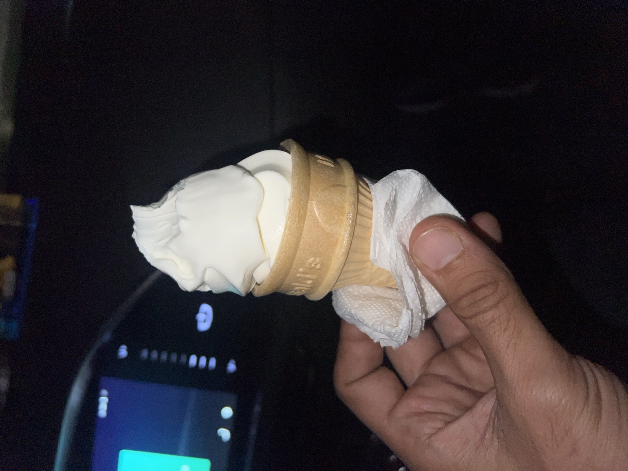 In the city of Sabunchu Raion, Azerbaijan, a person is holding a vanilla soft-serve ice cream cone. The ice cream is white and swirled, resting in a waffle cone. A white paper napkin is wrapped around the base of the cone. The background is dark, with the illuminated dashboard of a car visible on the left side of the image. The overall setting suggests the image was taken at night, possibly inside a vehicle.