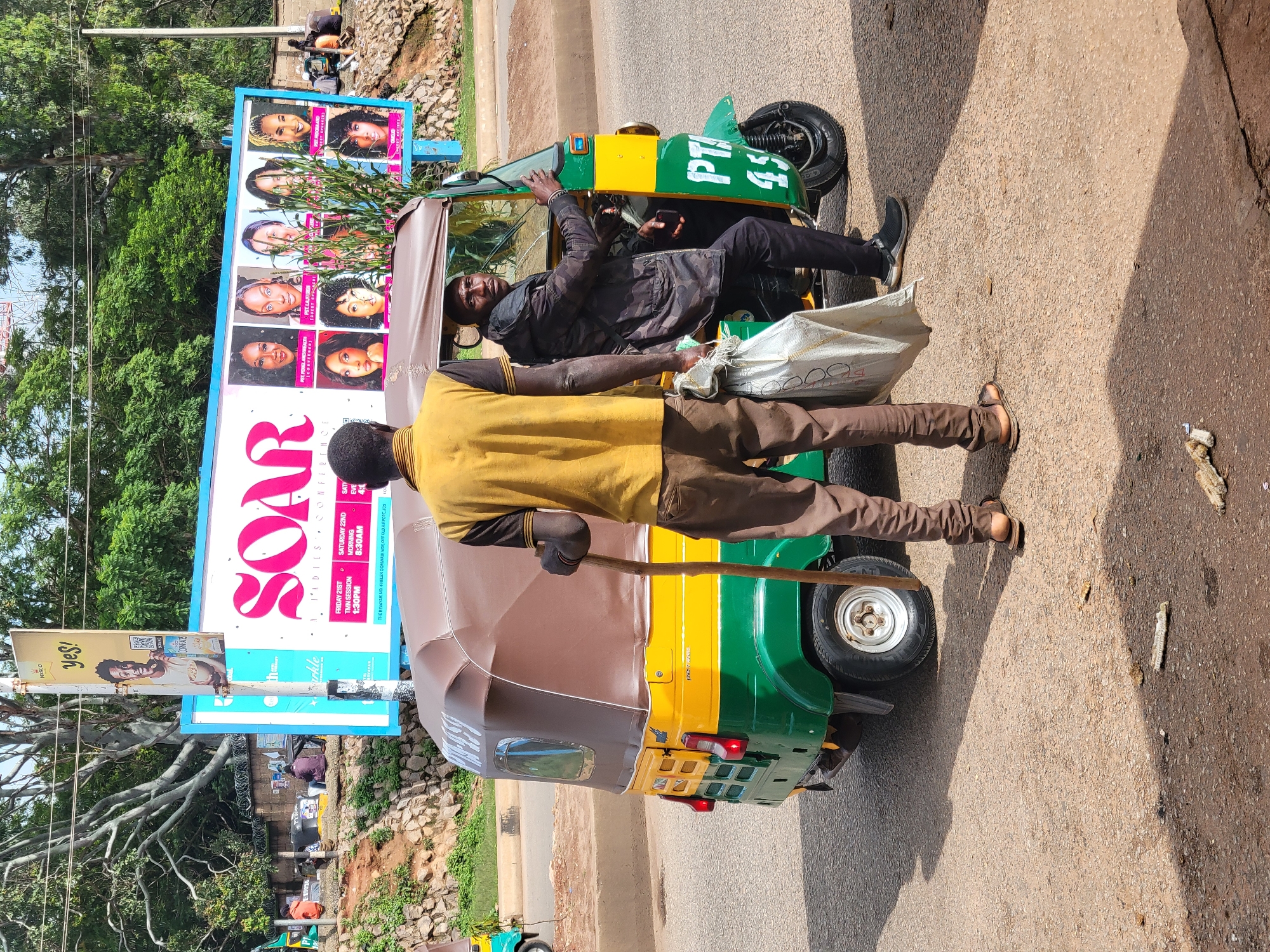 The image depicts a street scene in Jos, Nigeria, with a yellow and green auto-rickshaw positioned in the foreground. Two men are present: one, wearing a yellow polo shirt and brown trousers, stands facing away from the viewer, holding a wooden stick and a white sack; the other, dressed in a camouflage jacket and black trousers, sits in the driver's seat of the auto-rickshaw. A large billboard in the background advertises a "SOAR LADIES CONFERENCE" with images of women. The environment appears to be a roadside with a dirt and asphalt surface, and some vegetation visible.
