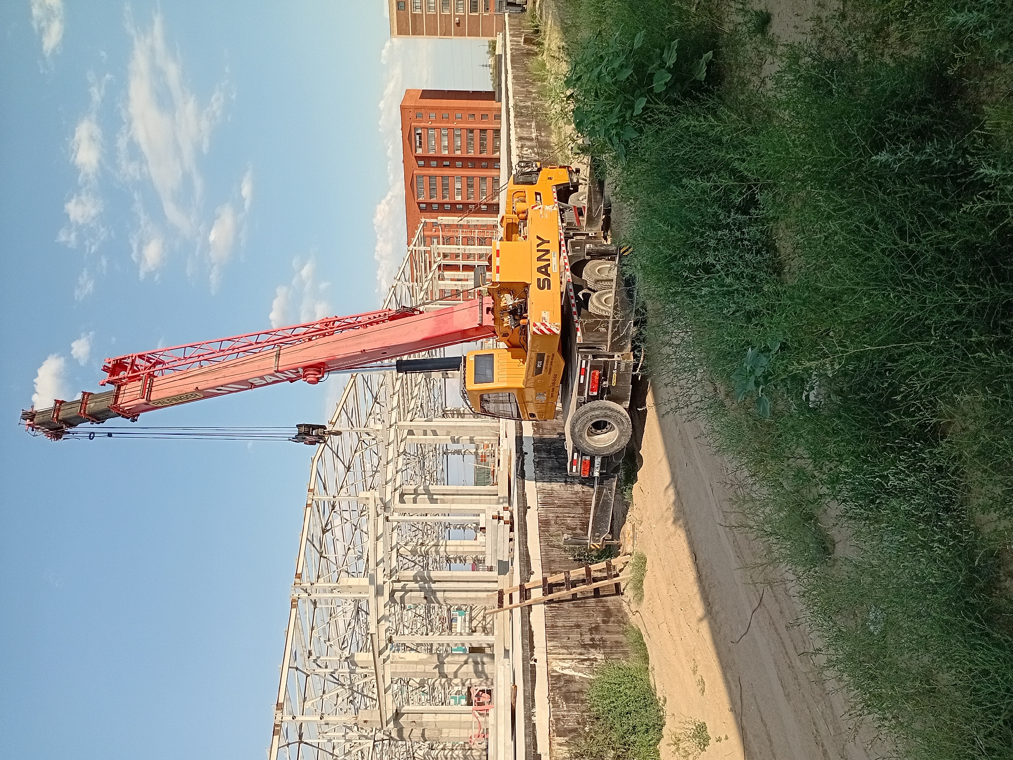 This is a medium-shot, eye-level, outdoor photograph taken on a sunny day, likely in the city of Semey, Kazakhstan, depicting a construction site. A large, red and yellow SANY mobile crane is positioned on a dirt path in the foreground. The crane's boom is extended upwards and to the left, with its hook assembly visible. To the left of the crane, a partially constructed building with a white steel framework stands prominently. This building appears to be undergoing assembly, with concrete pillars supporting the structure. A wooden ladder leans against a raised concrete platform in front of the building. In the background, two multi-story brick residential buildings are visible, one orange-red and the other a lighter brown. The ground is a mix of sand and sparse green vegetation. The sky is blue with scattered white clouds. There are no people visible in the frame.
