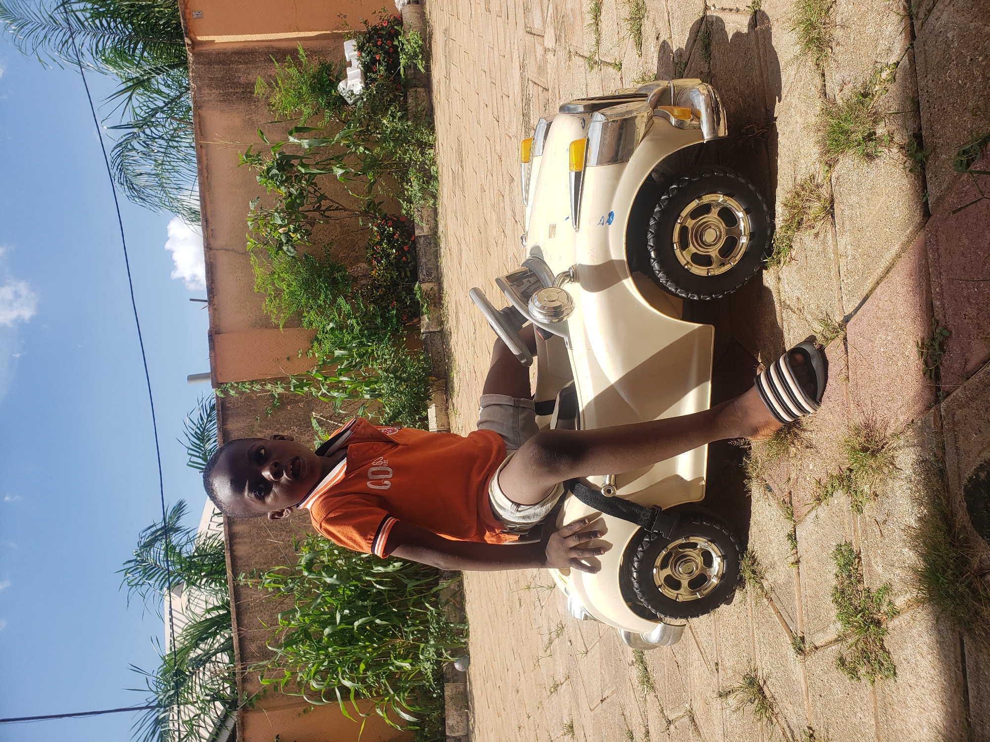 A young boy is sitting in a cream-colored toy car. He is wearing an orange polo shirt with white and black trim and light-colored shorts. The boy is looking towards the camera with his mouth slightly open. He is wearing black and white striped sandals. The toy car has chrome accents, yellow headlights, and black tires with gold rims. The boy's left hand is resting on the steering wheel of the car. The scene appears to be outdoors, with a paved ground made of interlocking bricks. In the background, there is a tall, orange wall, with lush green plants and trees growing in front of it. A bright blue sky with scattered white clouds is visible above. The overall setting suggests a residential area in Sabon Tasha, Nigeria, possibly a backyard or driveway.