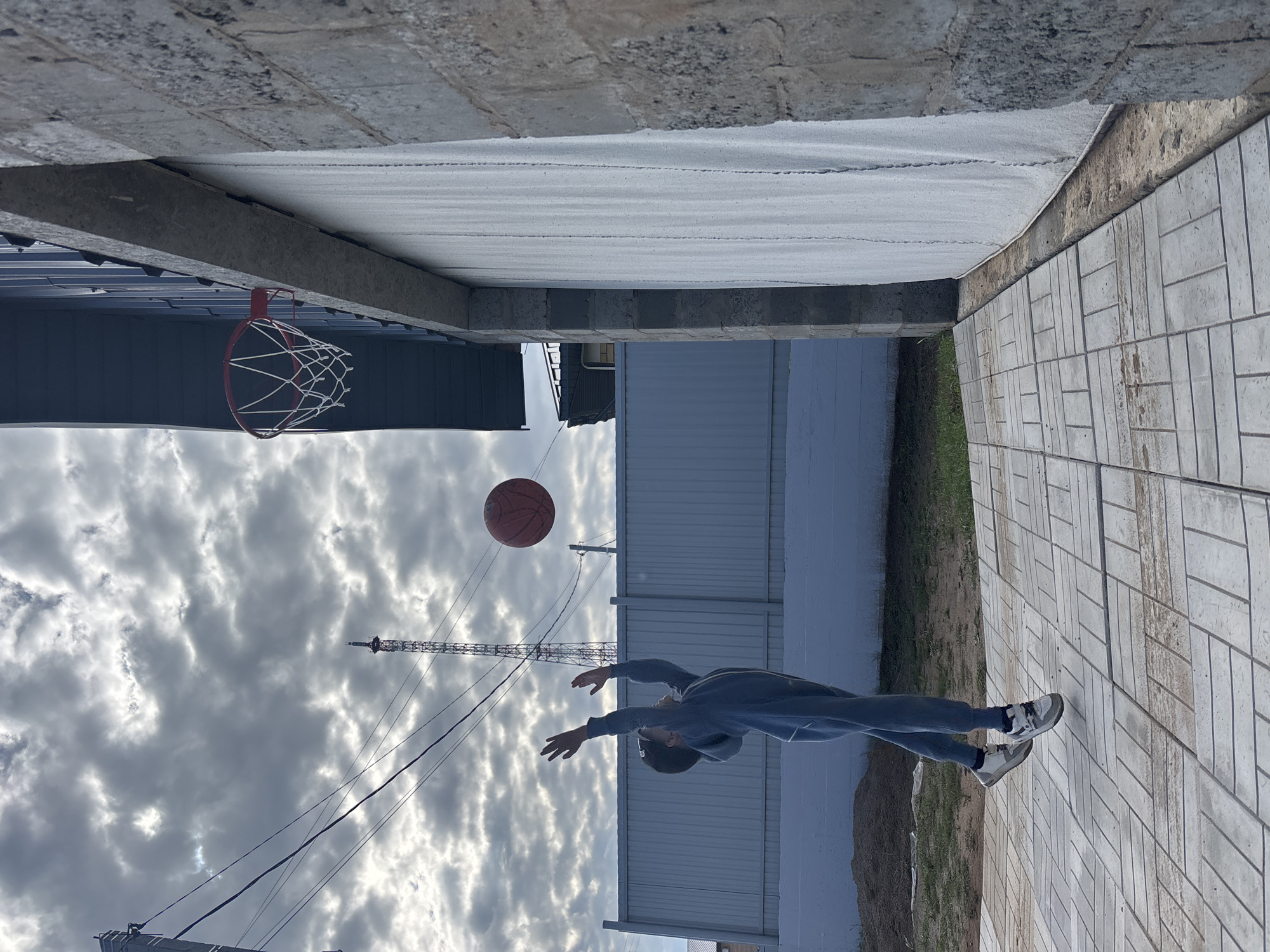 An outdoor scene in Chaikovsky, Russia, captures a dynamic moment of a young person playing basketball. The central subject is a person, likely male, dressed in a blue hoodie and jeans with sneakers, reaching upwards with both hands towards an orange basketball suspended in mid-air. To the left, a red basketball hoop with a white net is attached to an overhead concrete structure, suggesting a makeshift or informal playing area.
The ground is paved with light grey rectangular stones arranged in a patterned layout. In the background, various buildings with blue/grey corrugated metal siding are visible. A tall, lattice-like tower, possibly a communication tower or crane, stands prominently against a cloudy, overcast sky. The overhead concrete structure under which the game is taking place suggests the location might be under a bridge, overpass, or a similar architectural feature. The lighting indicates it is daytime, but the heavy cloud cover prevents direct sunlight. The overall atmosphere is one of focused activity in a functional, possibly industrial-adjacent, urban setting. There is no visible text in the image.