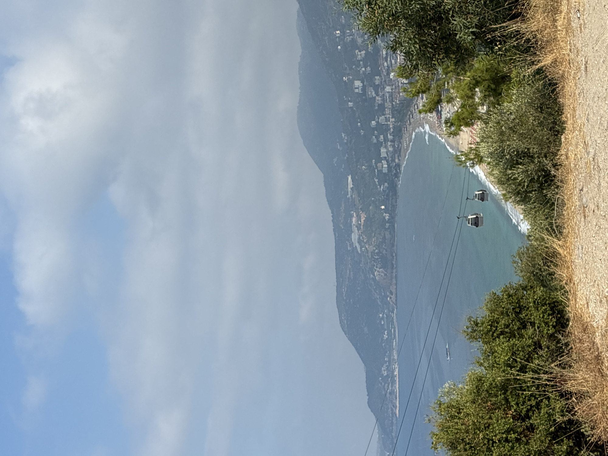This is a medium shot, taken from an elevated perspective, of the coastline of Alanya, Turkey. In the foreground, there is dry, tall grass and a gravelly surface. A lush green tree is on the right side of the frame. The middle ground features a sandy beach with some umbrellas and buildings lining its edge. The sea, with a boat in the distance, is a bright blue and appears calm. Two gondolas are visible suspended from cables over the sea. In the background, a large, green, hilly landscape rises, dotted with buildings. The sky is a hazy blue with scattered clouds.