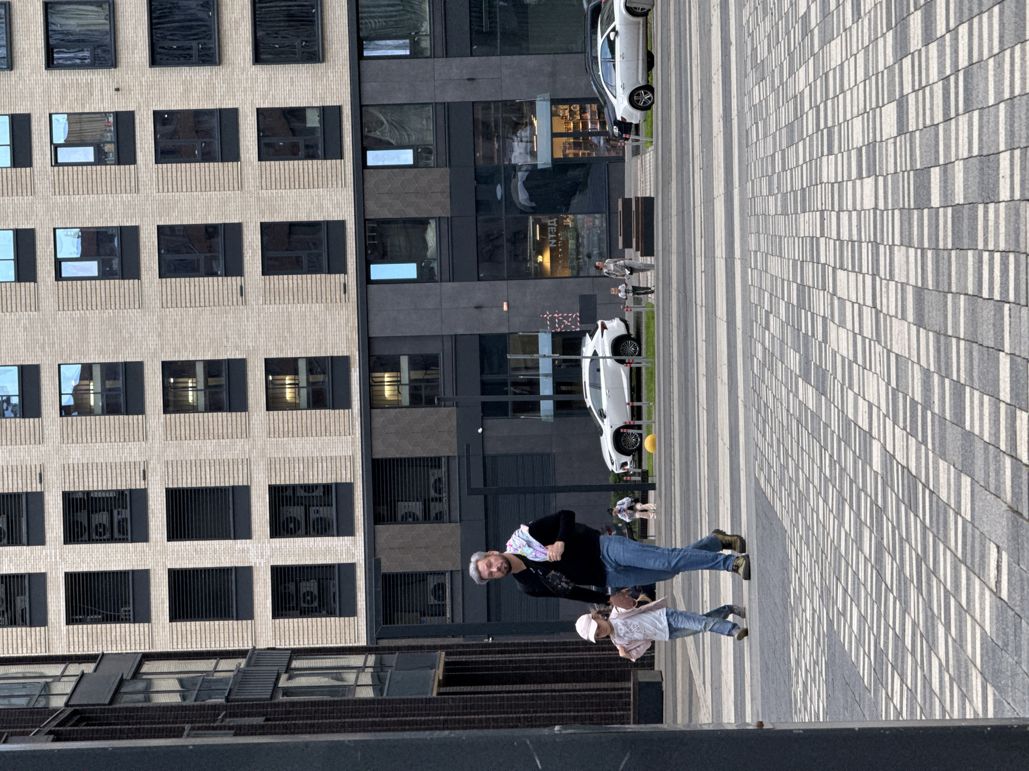 A man and a young girl walk on a sun-drenched paved plaza in front of a modern building. The man, wearing a black shirt and blue jeans, has gray hair and is looking towards the camera. He has a colorful scarf draped over his shoulders. The girl, wearing a light pink top, a white cap, and blue jeans, walks beside him, holding his hand. The paved ground is a mosaic of light and dark gray rectangular tiles, creating a striped pattern with alternating shadows cast by an unseen object.
The building behind them is a multi-story structure with a facade of light beige brick on the left and dark gray panels on the right. The right side of the building has large windows, some reflecting the sky. Several white cars are parked in the background, including a white sedan further up the plaza and another white vehicle that appears to be an SUV partially visible near the man and girl.
The scene suggests a pleasant day, likely afternoon, given the strong shadows. The overall atmosphere is casual and urban. There is no visible text in the image.