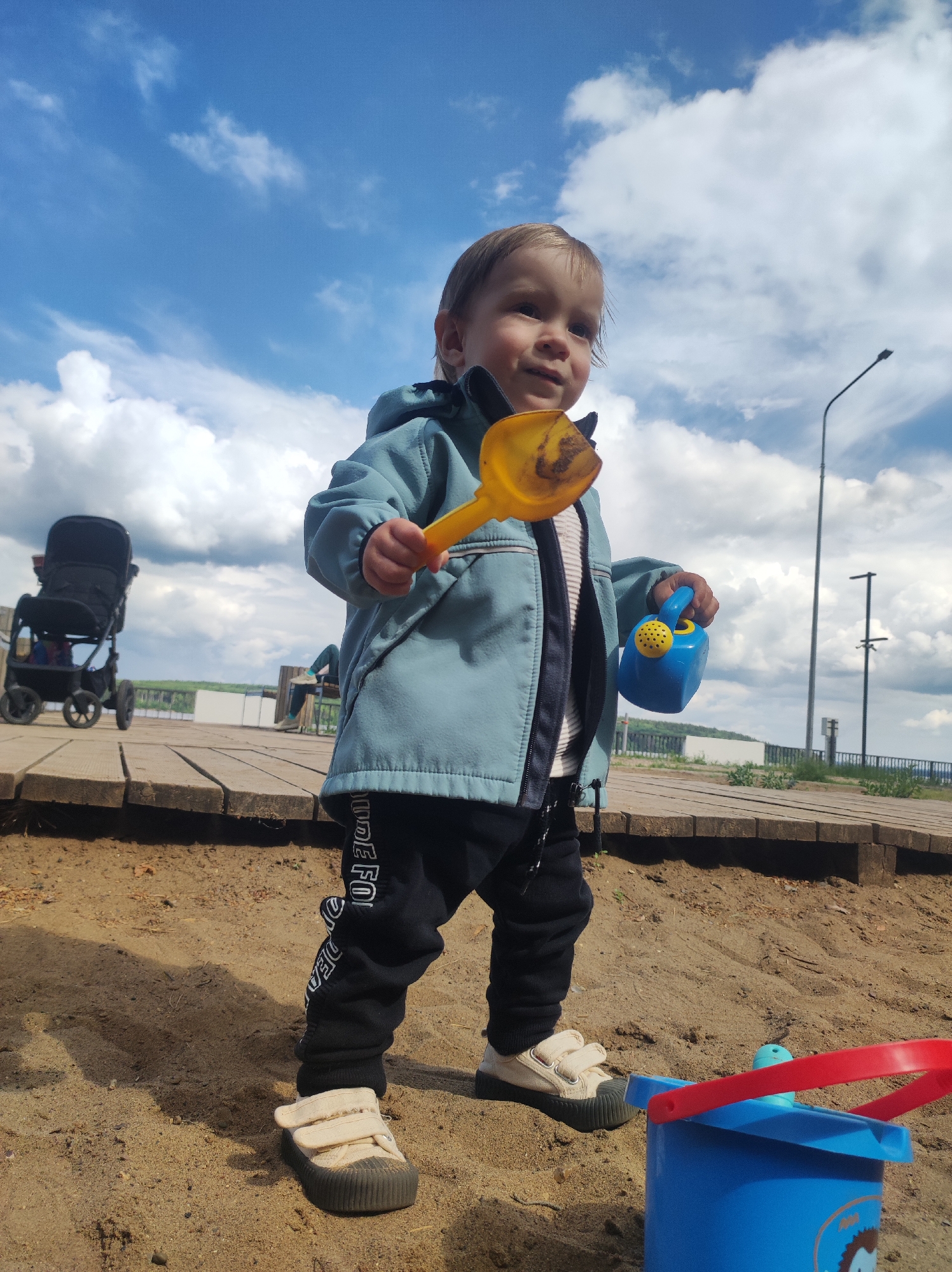 A toddler stands in a sandbox, holding a yellow sand shovel in their left hand and a blue watering can in their right. The toddler wears a light blue jacket, dark-colored pants with white lettering, and light-colored shoes. A blue bucket with a red handle sits in the sand near the toddler's feet.
The sandbox is situated near a wooden platform. In the background, a black stroller sits on the platform. Further behind, there is a railing, a grassy area, and a body of water. Light posts stand against a backdrop of a blue sky with scattered clouds.
The image was taken in Chaikovsky, Russia.