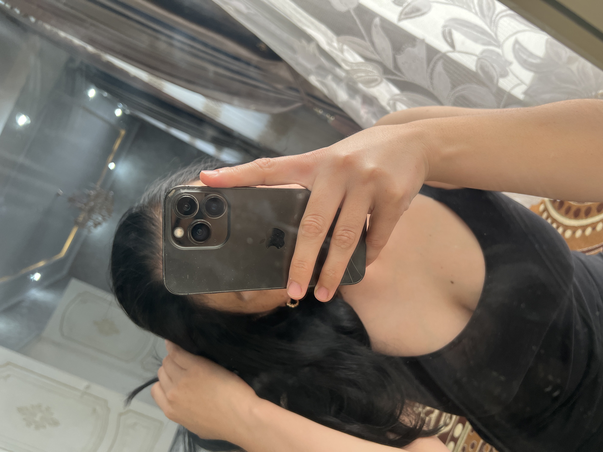 A person is taking a mirror selfie in a room. The person is holding a smartphone in their right hand, angled to capture their reflection. They have long, dark hair and are wearing a black tank top. The background shows a large mirror reflecting the room, which includes patterned wallpaper on the ceiling, curtains, and what appears to be a chandelier or light fixture. The floor has a patterned rug. The location is identified as Kara-Dıykan, Kyrgyzstan.