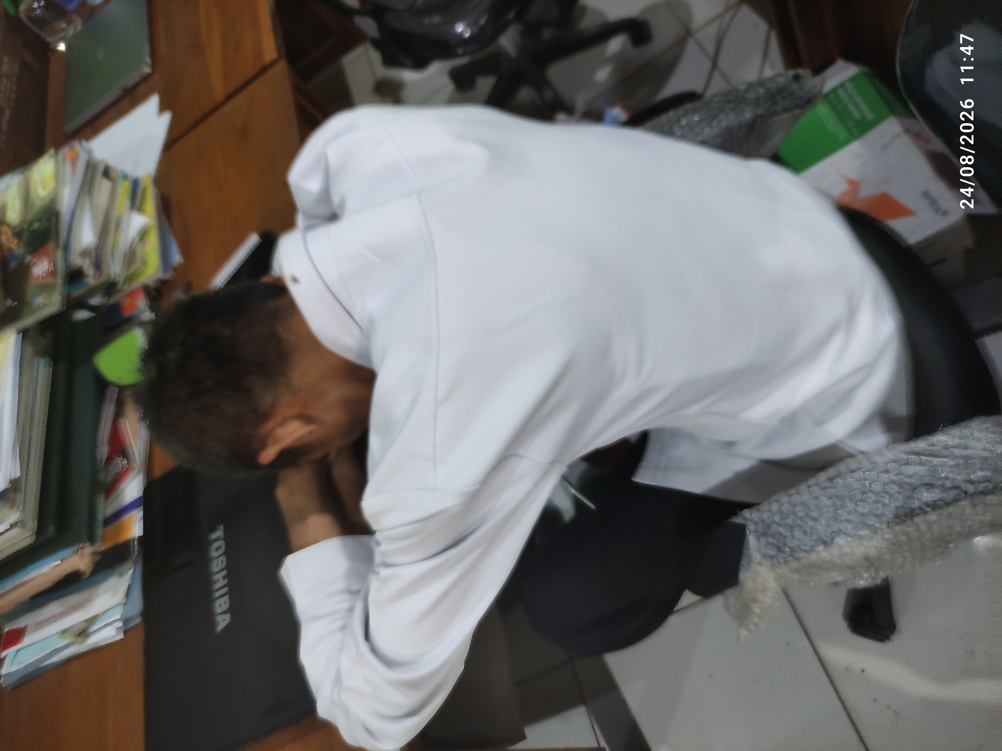 An overhead, slightly angled, medium shot captures an individual resting their head on a dark-colored laptop, inscribed with the brand "TOSHIBA," while seated at a wooden desk. The person is wearing a white long-sleeved shirt and dark pants. Their arms are crossed, and their head is bowed, suggesting they are asleep or resting. The desk is cluttered with various books and papers. To the right of the frame, a chair with a black seat and chrome legs is partially visible, along with a green and orange box on the floor. The background shows a tiled floor and a hint of a wall. The timestamp "24/08/2026 11:47" is present in the bottom right corner of the image. The scene appears to be indoors, possibly an office or study area in Tamanrejo, Indonesia.