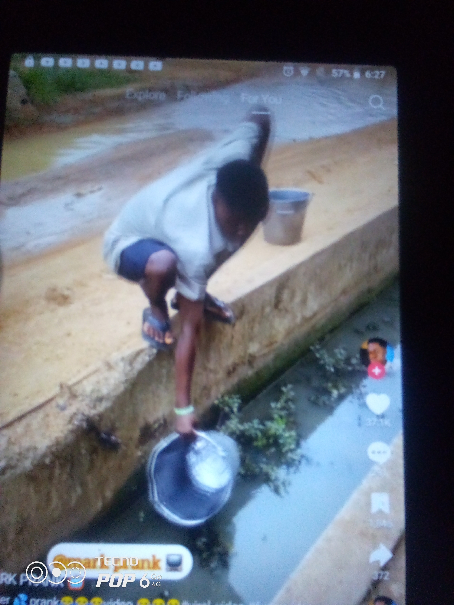 A person is squatting on a raised concrete ledge next to a narrow, muddy channel filled with murky water and some green vegetation. They are wearing a light-colored t-shirt, dark shorts, and flip-flops. The person is reaching down with their right hand, holding a silver bucket that appears to be partially submerged in the channel, and is possibly collecting water. A second, gray bucket is visible on the ledge behind them. The environment appears to be an outdoor, possibly semi-urban or rural area, with a dirt path and a wall in the background. The image is taken from a slightly elevated angle and appears to be a screenshot from a mobile device, indicated by UI elements at the top and side of the screen, including icons for battery life, Wi-Fi, and what seem to be social media interaction buttons. The exact location in Howayi, Nigeria, cannot be determined from the visual information present.