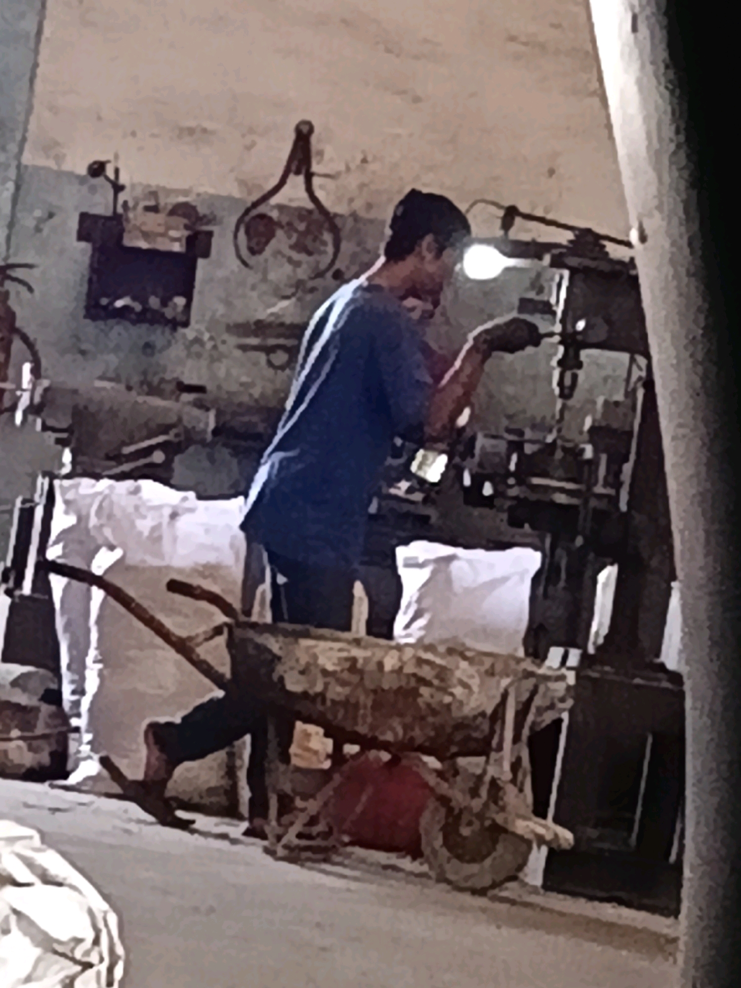 In a dimly lit workshop, a man in a blue t-shirt is operating a piece of machinery. He is wearing dark pants and gloves, and a black cap. The machine, which appears to be a drill press or similar industrial equipment, is illuminated by a bright overhead lamp. In the foreground, a rusty wheelbarrow filled with what appears to be wood or stone is positioned on the concrete floor. Behind the man and the machinery, there are stacks of white sacks and various tools or equipment mounted on the wall. The overall environment suggests an active workspace, possibly a metal fabrication or repair shop. This scene is located in Belimbing, Indonesia.