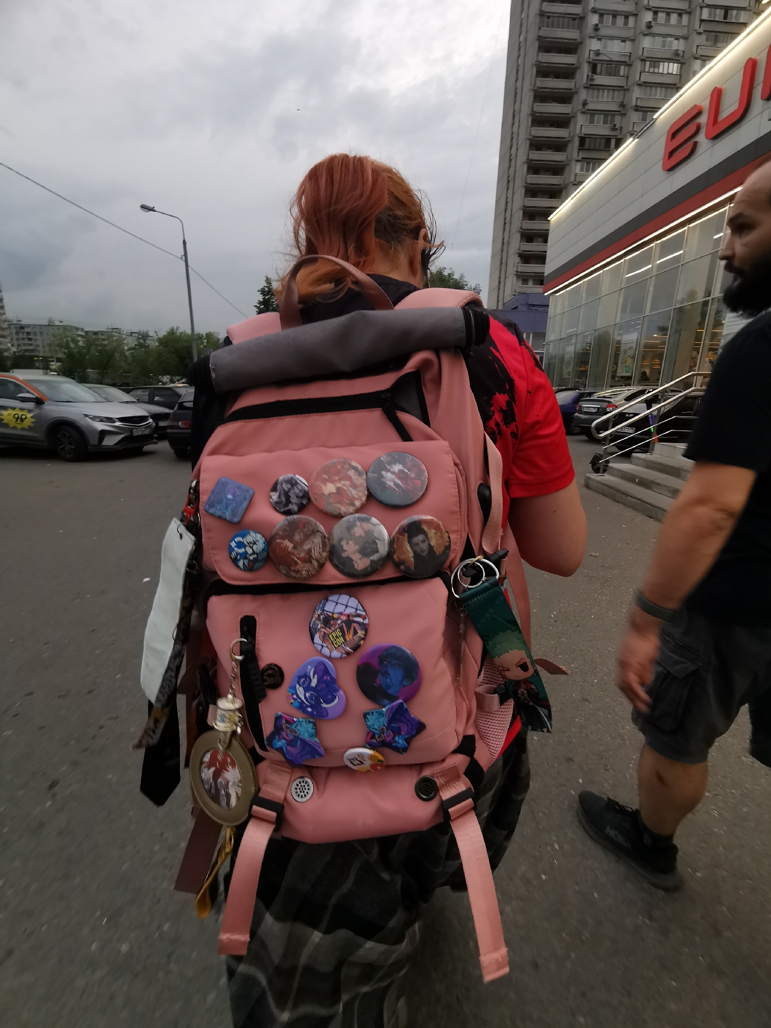 A street scene in an urban environment of Unknown, Unknown, features a person with reddish-orange hair, wearing a red and black shirt, and carrying a pink backpack adorned with multiple buttons and trinkets. The backpack also has several straps and tassels hanging from it. The person is walking away from the camera, slightly angled to the left. To the right of the person is another individual partially visible, wearing a black shirt and grey shorts.
In the background, cars are parked on the street, including a silver taxi. A large building with a sign that reads "EURO" is visible. The sky is overcast and grey.