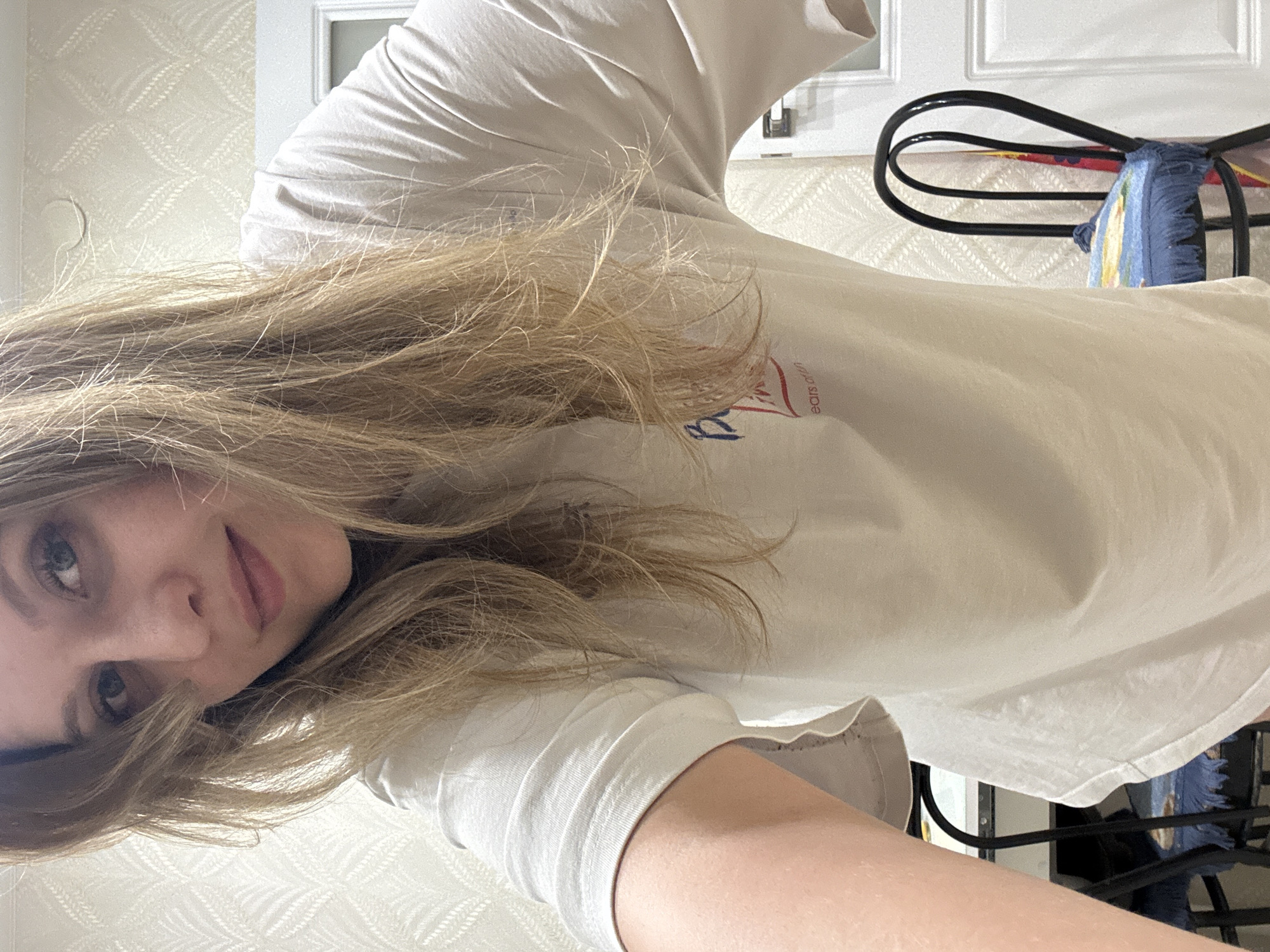 A young woman with long, blonde hair is wearing a light-colored t-shirt, posing for a selfie indoors. She is fair-skinned with light-colored eyes. Behind her, there is a white door and wallpaper with a repeating pattern. Part of a black chair with a blue cushion is visible in the background. No information suggests the location is Izhevsk, Russia.