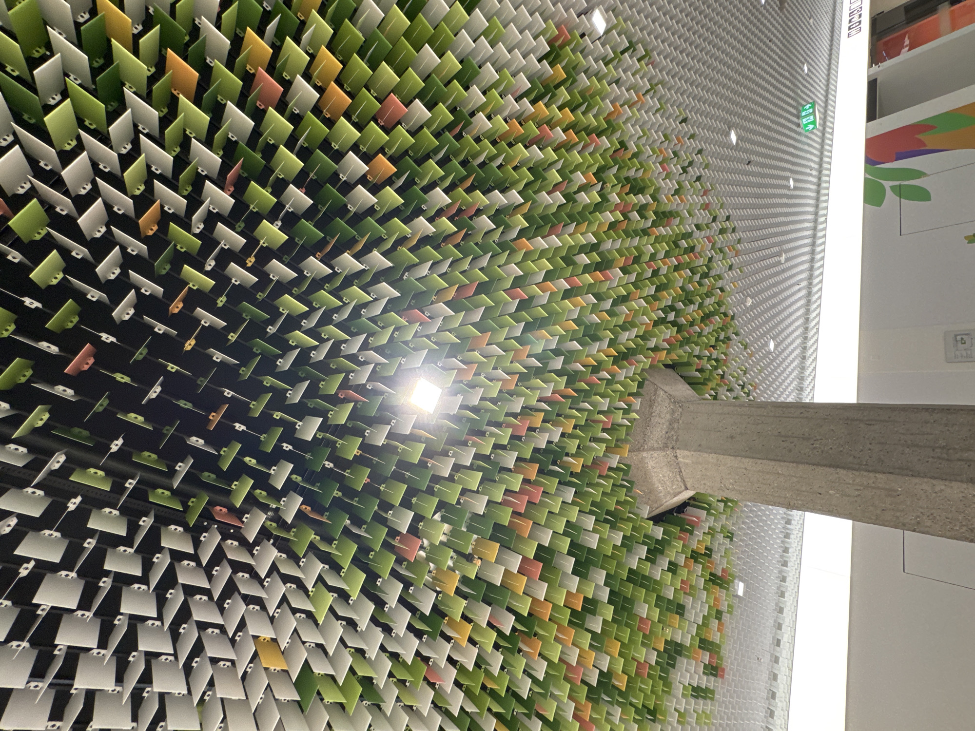 The image shows a ceiling designed to resemble a canopy of leaves. It is composed of numerous small, rectangular panels in shades of green, white, orange, and yellow, attached to a dark ceiling. The panels are angled and layered, creating a textured, organic effect. A large, concrete pillar, textured like rough stone, extends upwards from below, supporting the ceiling structure and appearing as a tree trunk. The background shows a white ceiling with a grid pattern and some lighting fixtures. There is a green exit sign visible in the upper right portion of the image, indicating an indoor location. The overall impression is a modern, artistic installation, likely in a public space. The location is identified as Morbio Inferiore, Switzerland. There are no people visible in the image.