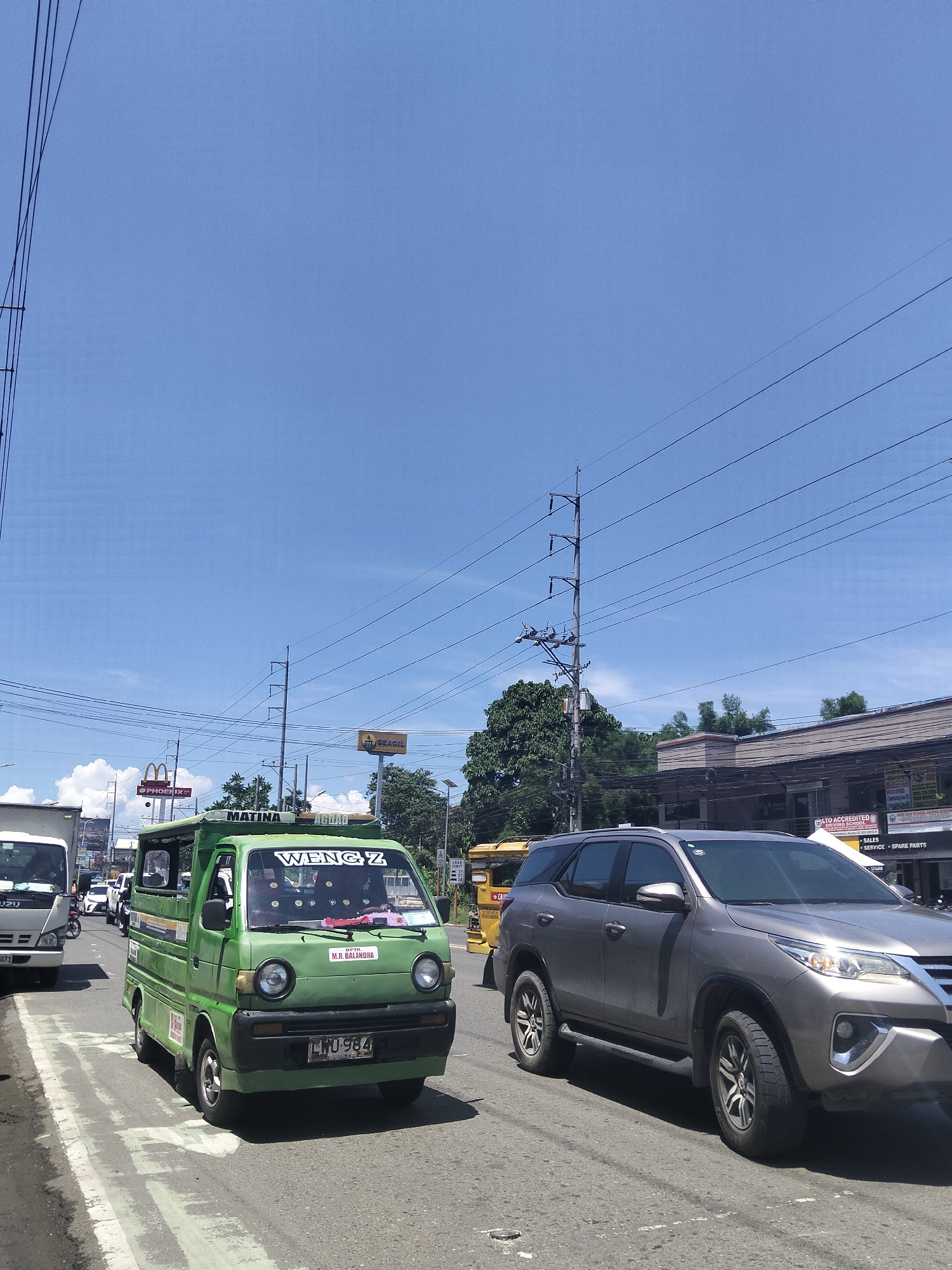 This is a daytime, outdoor, eye-level shot of a street scene in Davao City, Philippines. The sky is a clear, bright blue with some scattered clouds. Power lines crisscross the upper portion of the image.
The foreground is dominated by traffic. A green utility van with the name "WENGZ" and "MATINA" visible on its exterior is in the left lane. It has a license plate that reads "LWU-984". To the right of the green van is a silver SUV. Further back, a white truck is visible in the far left lane, and a yellow tricycle is in the right lane.
Several commercial establishments are visible on the right side of the street, including one with signage indicating it is an "LTO ACCREDITED DRIVING SCHOOL" offering "SALES, SERVICE, SPARE PARTS". A "SEAOIL" gas station sign is also visible, as is a "PHOENIX" sign and a McDonald's logo. The presence of these businesses and the style of the vehicles suggest an urban or suburban setting in the Philippines. The overall impression is of a busy, sunny day on a street with moderate traffic.