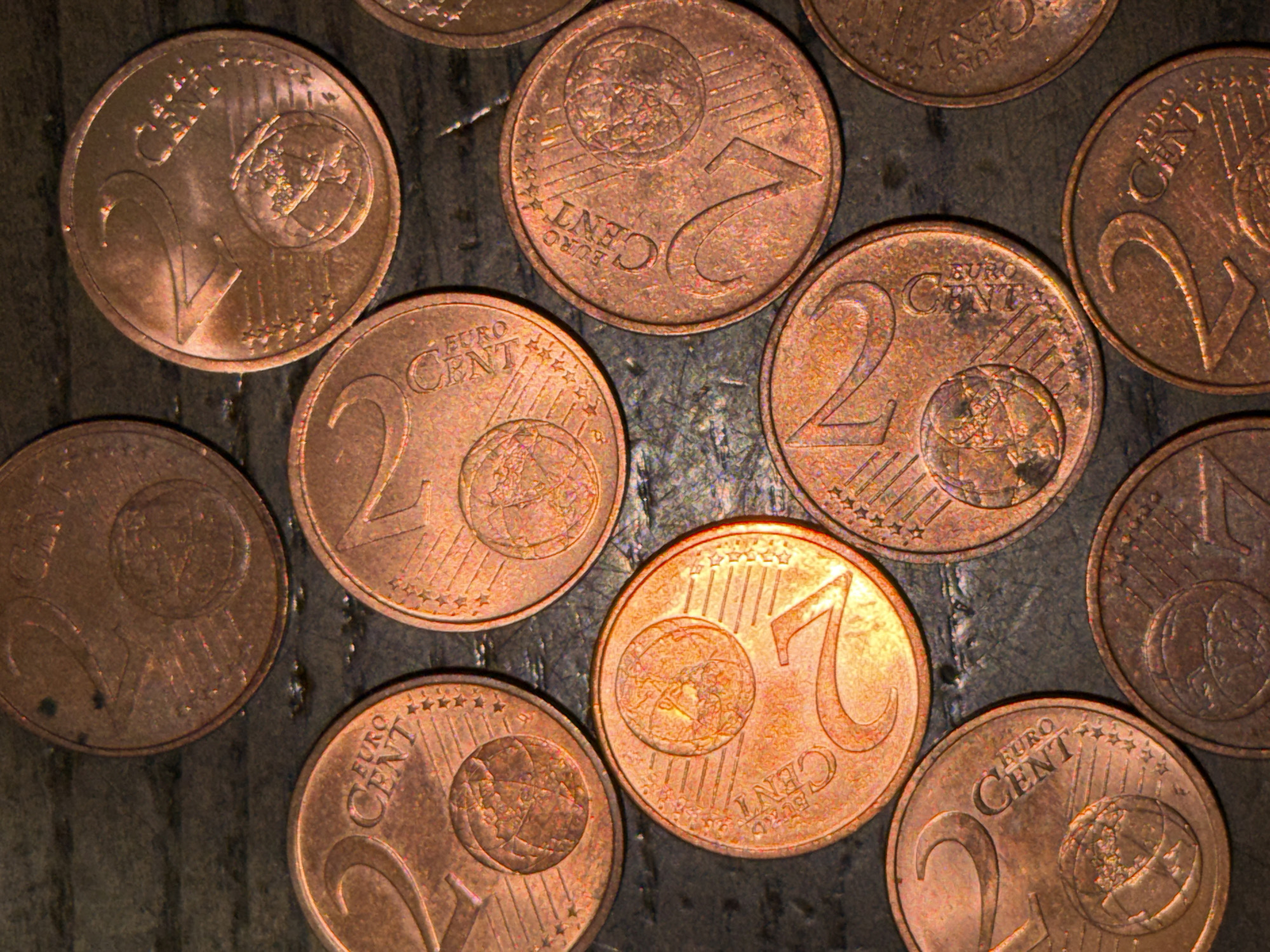 The image presents an overhead view of eleven 2-euro cent coins scattered across a dark, textured surface. Each coin is copper-colored and clearly displays the numeral "2" along with the text "EURO CENT" and the common Eurozone design featuring a globe of Europe, twelve stars, and six parallel lines. The coins are randomly oriented and positioned, with some slightly overlapping others. The underlying surface appears to be a dark, somewhat irregular or rough material, possibly wood or stone, providing a matte background to the metallic coins. The scene is observed in Strasbourg, France.
