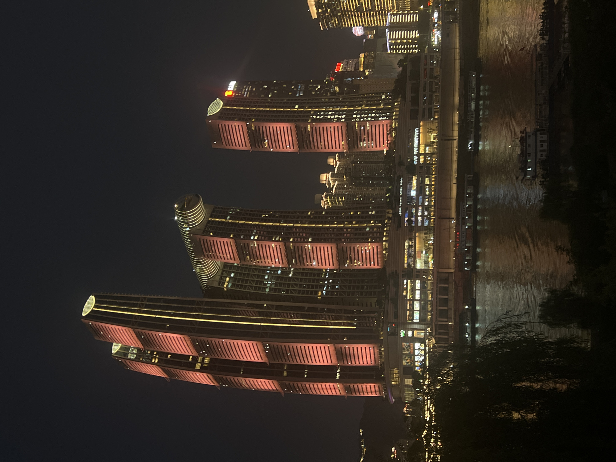 An illuminated city skyline at night is shown, with the Yangtze River flowing in the foreground. In the center of the frame, three prominent skyscrapers stand tall, their facades bathed in vibrant red and yellow lights. The tallest skyscraper on the left has a distinct circular top with yellow lights, while the building to its right features a large, curved architectural element. The third skyscraper, to the right, is a more slender rectangular building with red lighting on its facade. Below these main structures, a multi-story building complex is lit up with various lights, indicating commercial or residential use. Several boats are visible on the dark, reflective water of the river, with a larger ferry-like vessel in the lower right quadrant of the image carrying numerous passengers. On the far bank, a dense cluster of smaller buildings, also illuminated, stretches towards the horizon, with distant hills faintly visible in the dark sky. The foreground is dominated by the dark silhouettes of trees and foliage, including a weeping willow on the left, casting shadows over the scene. The overall impression is of a bustling, modern metropolis at night, with the river serving as a reflective artery through the urban landscape.