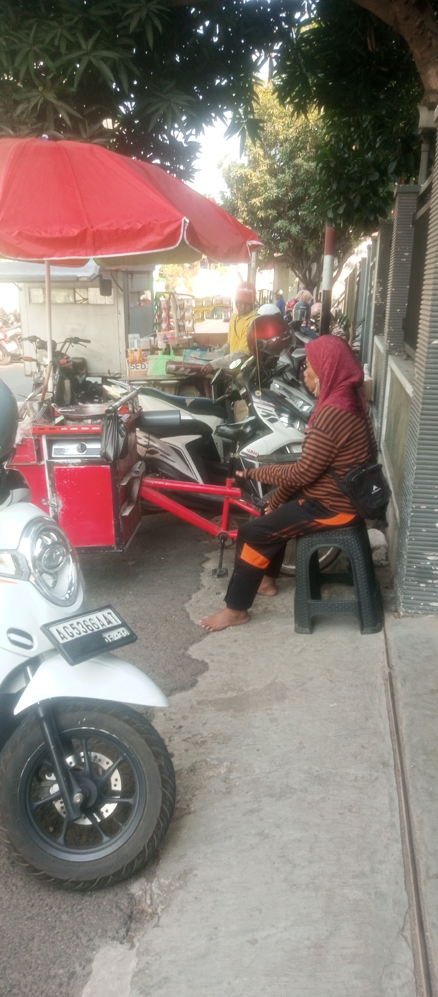 In the city of Kediri, Indonesia, a street vendor operates a small roadside stall under a large red umbrella, selling various items from a cart. Several motorcycles are parked around the stall, with one white scooter prominently positioned in the foreground with a license plate reading "AG 5366 AAT". A woman wearing a red and black striped long-sleeved shirt and black pants with orange accents sits on a black plastic stool, facing away from the camera, with her bare feet touching the ground. She has a black bag slung over her right shoulder. Behind her, another person in a yellow shirt and red head covering can be seen behind the stall. The environment appears to be a street with a paved walkway and a building with a ribbed facade to the right. Trees with green foliage provide shade overhead.