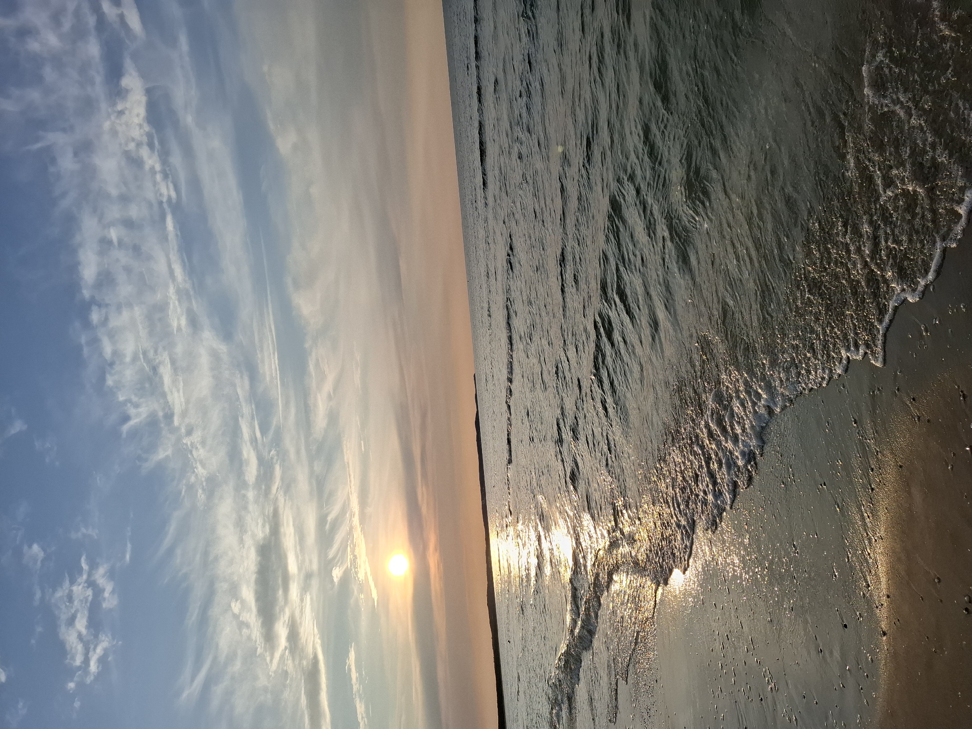The image depicts a serene beach scene at sunset in Świnoujście, Poland. The sun, a bright orb, is low on the horizon, casting a warm glow across the sky and reflecting on the gentle waves of the Baltic Sea. The sky is a canvas of soft blues and pastel oranges, with wispy cirrus clouds streaked across it, hinting at the approaching twilight.
The foreground is dominated by a sandy beach, wet from the receding tide, with small pebbles scattered across its surface. A small wave is breaking at the edge of the water, its white foam glistening in the sunlight. The sea itself is a deep blue-green, with ripples indicating a calm, steady breeze. In the distance, a dark, low-lying landmass, likely the coastline or a peninsula, is visible.
There are no people or specific objects clearly identifiable in the frame, emphasizing the natural beauty and tranquility of the location. The overall atmosphere is peaceful and picturesque, characteristic of a coastal sunset.