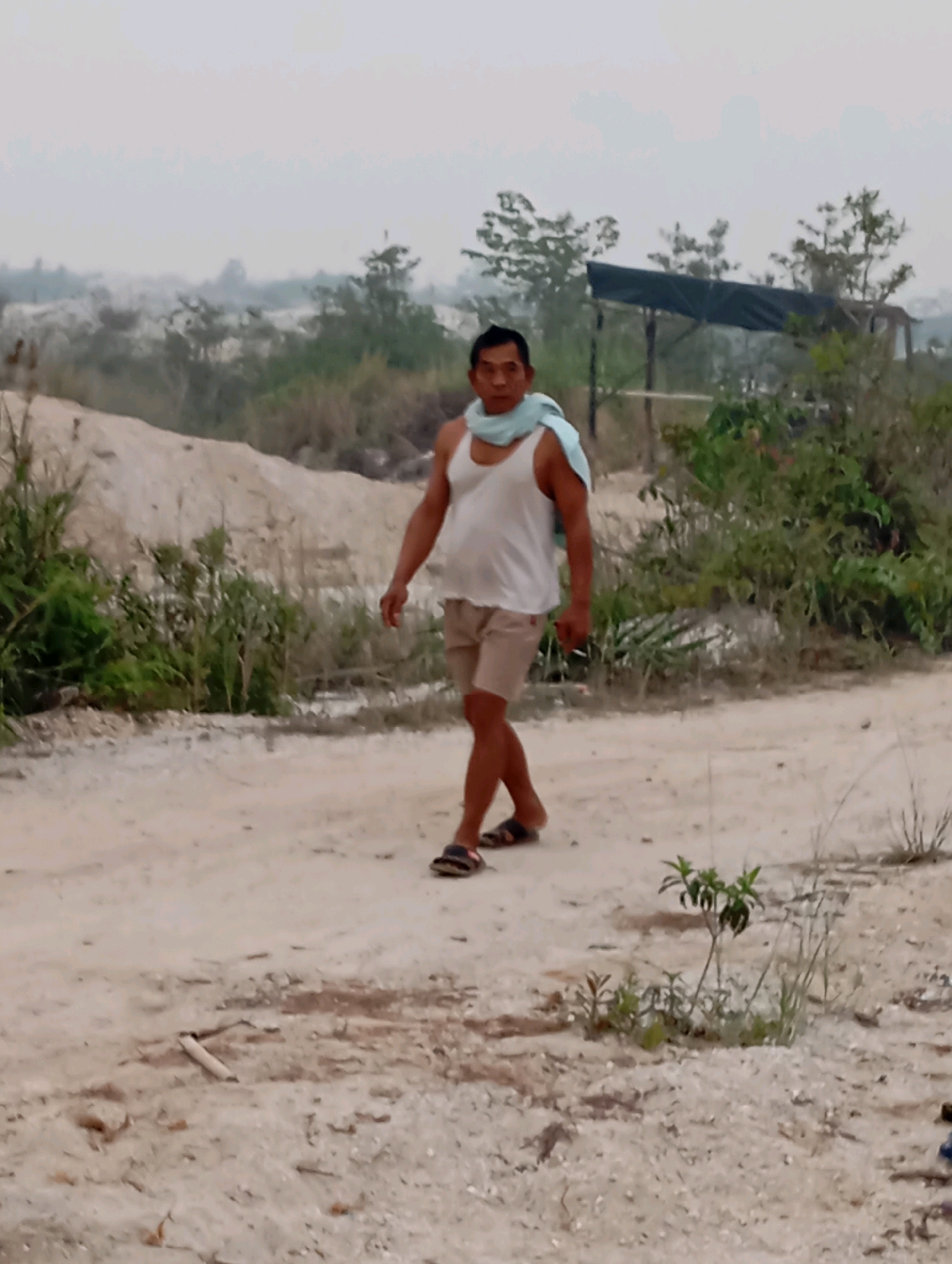 A middle-aged man is walking on a dirt road in Tumbang Tariak, Indonesia. He is wearing a white tank top, light-colored shorts, and sandals. A light blue scarf is draped around his neck and shoulders. The background shows a barren landscape with sparse vegetation, sand dunes, and a covered structure in the distance. The sky is overcast. The man appears to be walking towards the viewer.