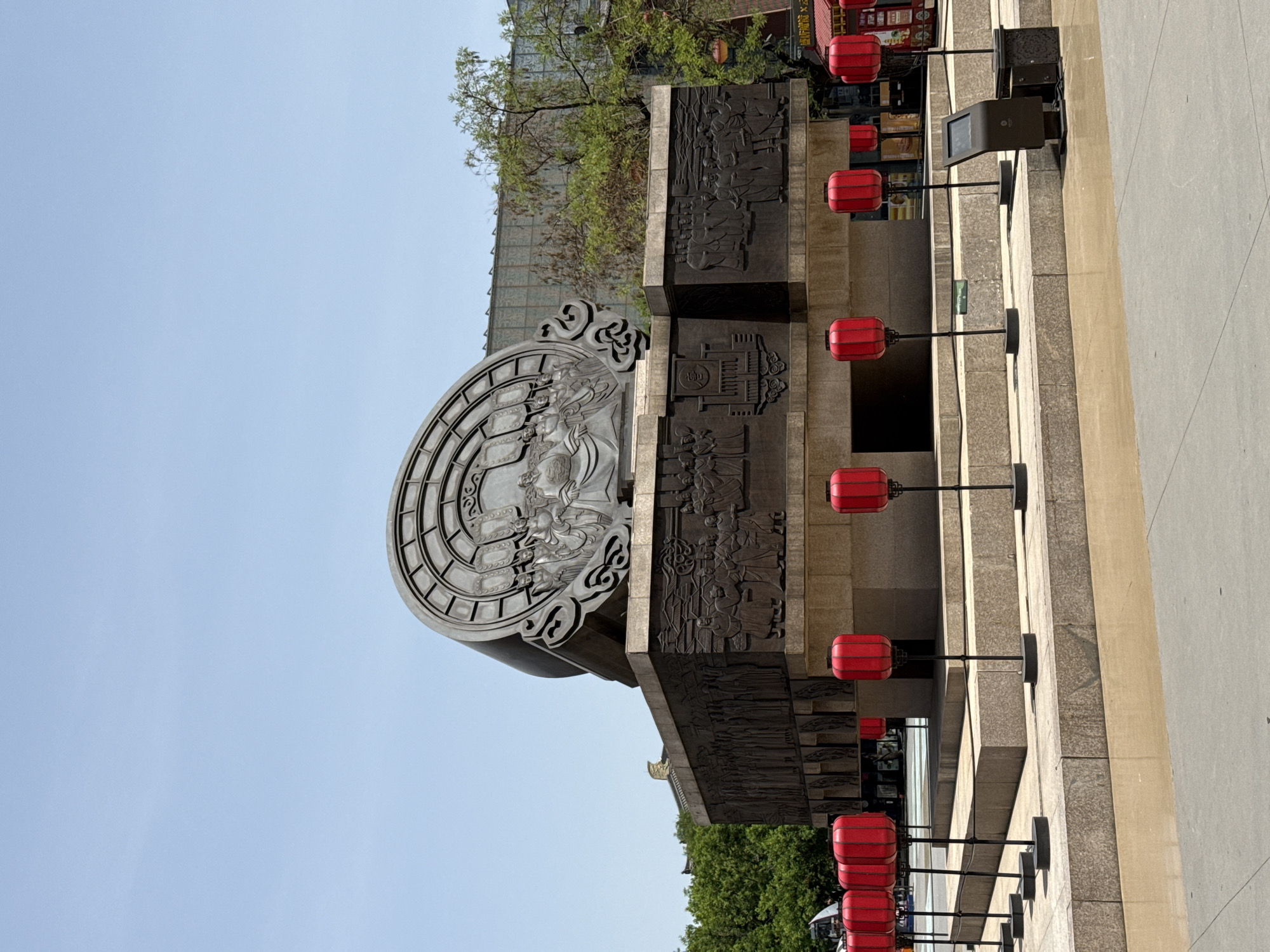 A low-angle, medium shot depicts a large, monumental structure in Yanta District, China. The structure is characterized by a semicircular, ornate relief with figures, positioned above a rectangular plinth adorned with bas-reliefs showing scenes from daily life and historical events. The relief artwork is detailed and possibly made of stone or bronze.
The building housing this structure is constructed of beige stone with modern lighting, a row of red lanterns hangs off to the right. Some green trees are seen at the top, and a blue sky is in the background.
