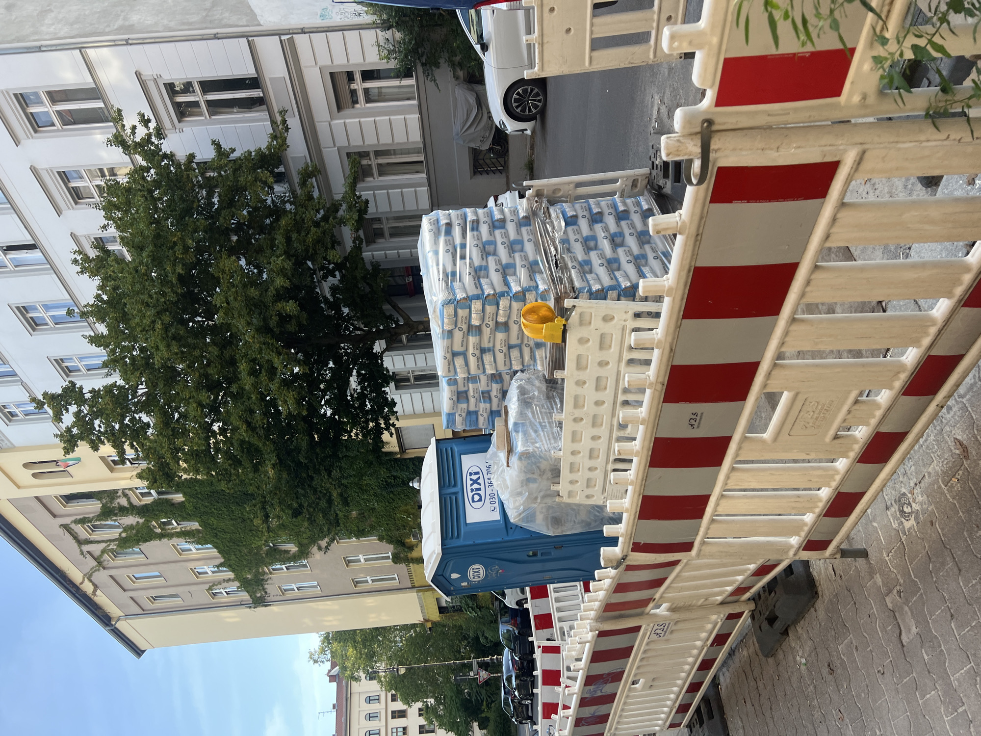 The image depicts a construction site on a city street in Berlin, Germany, likely in a residential or mixed-use area. In the foreground, a red and white temporary construction barrier delineates the work zone. Behind the barrier, a blue portable toilet with the "DIXI" logo is visible. Stacked high on the right are numerous bags of construction material, possibly cement or similar, wrapped in plastic. A yellow construction light is placed on top of one of the stacks.
The background reveals apartment buildings with classic European architecture, some adorned with ivy. Cars are parked along the street. The sky is a clear blue. The scene suggests ongoing construction work or road maintenance within the urban environment of Berlin. The street is paved with cobblestones in the immediate foreground, transitioning to asphalt further back.