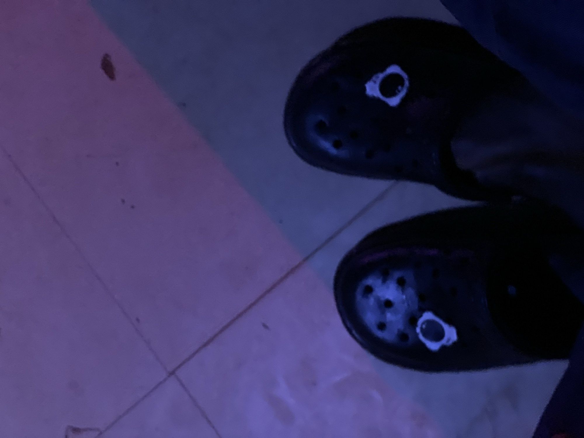 The image shows a close-up, overhead view of two black Crocs shoes on a tiled floor. The shoes are decorated with small, white Jibbitz charms that appear to be white circular designs with darker centers, resembling eyes. The floor tiles are light-colored and have visible grout lines, with a diagonal pattern formed by light and shadow, suggesting a light source from the right. The overall lighting is dim and has a strong purple hue, indicating artificial lighting, possibly from LED lights. There is no visible indication of the shoes' location in the city of Durumi, Nigeria.