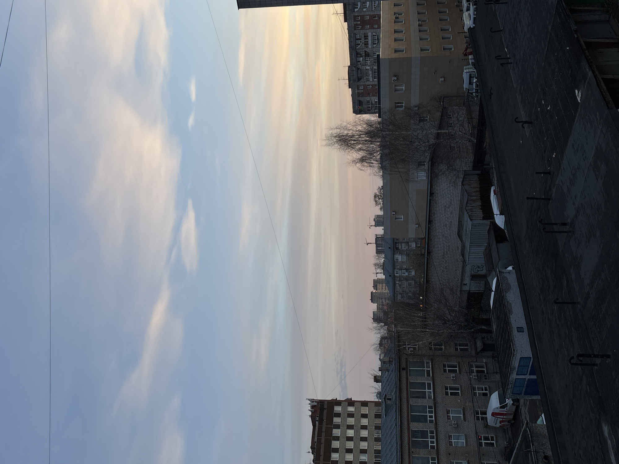 This image depicts a cityscape in Perm, Russia, observed from an elevated position, likely a rooftop or a high-story window. The focus is on a mix of buildings ranging from residential apartments to commercial or industrial structures. The sky occupies a significant portion of the frame, showing a transition of colors, possibly indicating sunrise or sunset. Several bare trees are scattered among the buildings, suggesting it might be late autumn or winter. A few vehicles are visible, and utility lines cut across the sky. The overall atmosphere is urban, with a mix of older and more modern construction styles.