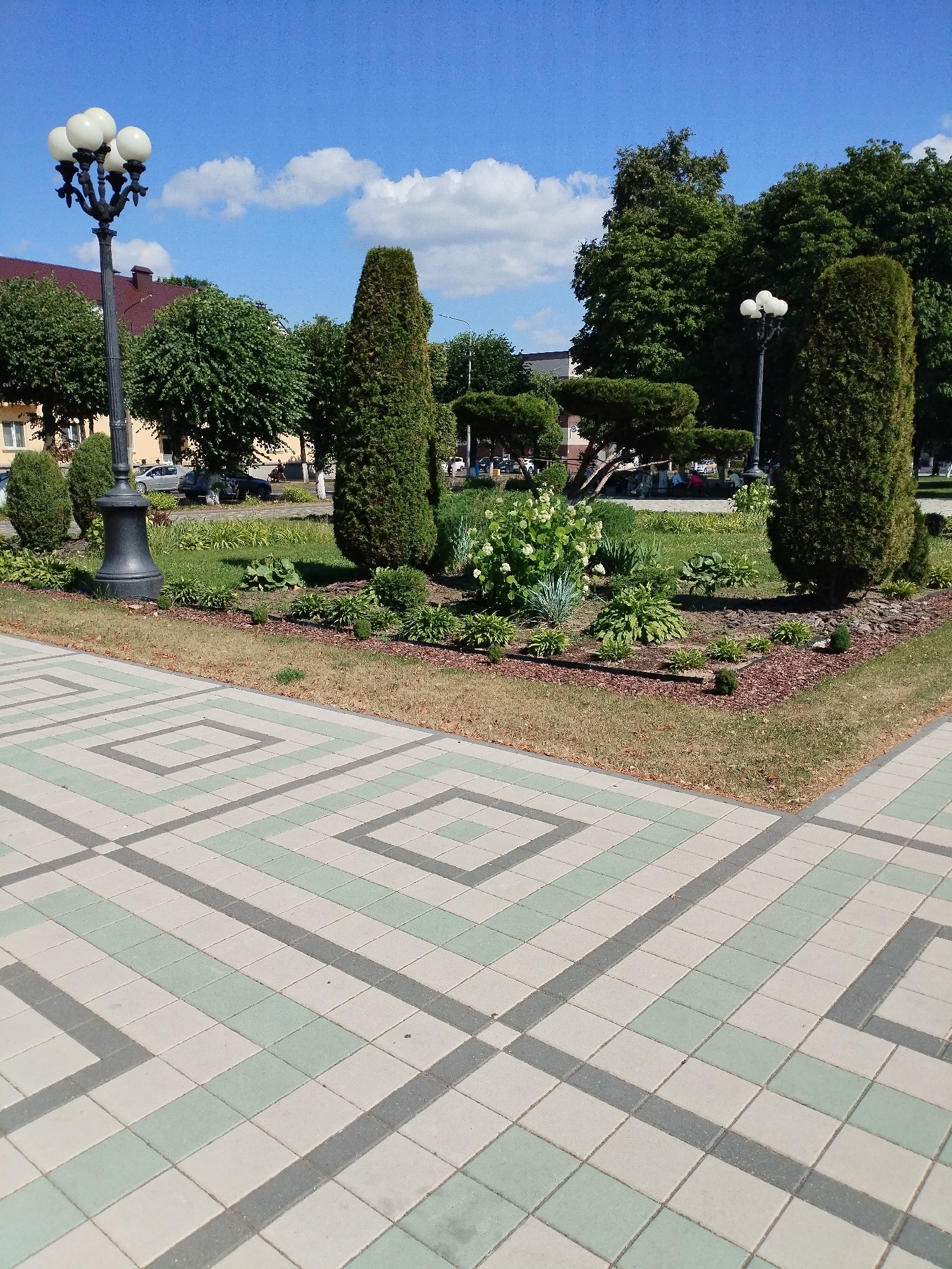 The image depicts a bright, sunny day in a park or public square, likely in Barysaw, Belarus. The foreground is dominated by a decorative paved walkway with a geometric pattern of light gray and pale green square tiles. Beyond the walkway, a landscaped garden area features various shrubs and plants, including a large cluster of white-flowering hydrangeas. Several tall, conical evergreen trees and rounded, manicured bushes are strategically placed within the garden. A lamppost with five globe-shaped lights stands to the left, while another similar lamppost is visible in the mid-ground to the right. In the background, a building with a reddish-brown roof and light-colored walls is partially visible, along with more trees and a glimpse of parked cars and a few people sitting on a bench. The sky is a vibrant blue with scattered white, puffy clouds, suggesting pleasant weather. The overall impression is one of a well-maintained, public outdoor space on a clear summer day. There is no visible text in the image.
