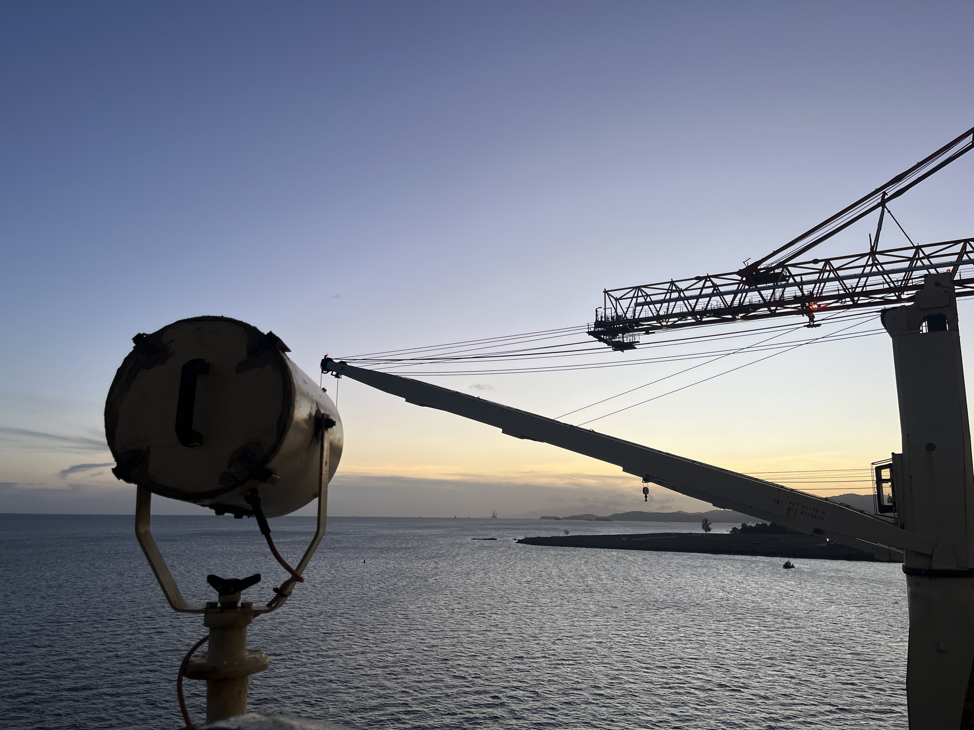 The image captures a serene marine industrial scene during either sunrise or sunset, characterized by warm light on the horizon and a cooling blue sky overhead. In the immediate foreground on the left, a large, cylindrical searchlight or spotlight is prominently featured, silhouetted against the fading light. It's mounted on a stand with visible red wiring.
Dominating the right and center of the frame is a massive industrial crane, likely belonging to a ship or port facility. Its intricate lattice boom structure is silhouetted against the sky, extending from the right edge towards the center-left. The crane's main arm stretches out over the water. Visible text on the crane arm indicates its specifications: "SWL 45 t 4.8-28m" (Safe Working Load 45 tonnes at a radius of 4.8-28 meters) and "40 t 4.8-36m" (40 tonnes at 4.8-36 meters).
The middle ground and background consist of a vast body of water, likely an ocean or large bay, showing gentle ripples reflecting the low light. In the distance, a dark coastline with hills or mountains is visible, along with some indistinct structures. Several small, distant boats or ships are dotted across the water, appearing as faint silhouettes. The overall atmosphere is tranquil, blending industrial elements with the natural beauty of the marine environment at twilight. No people or active events are discernible.