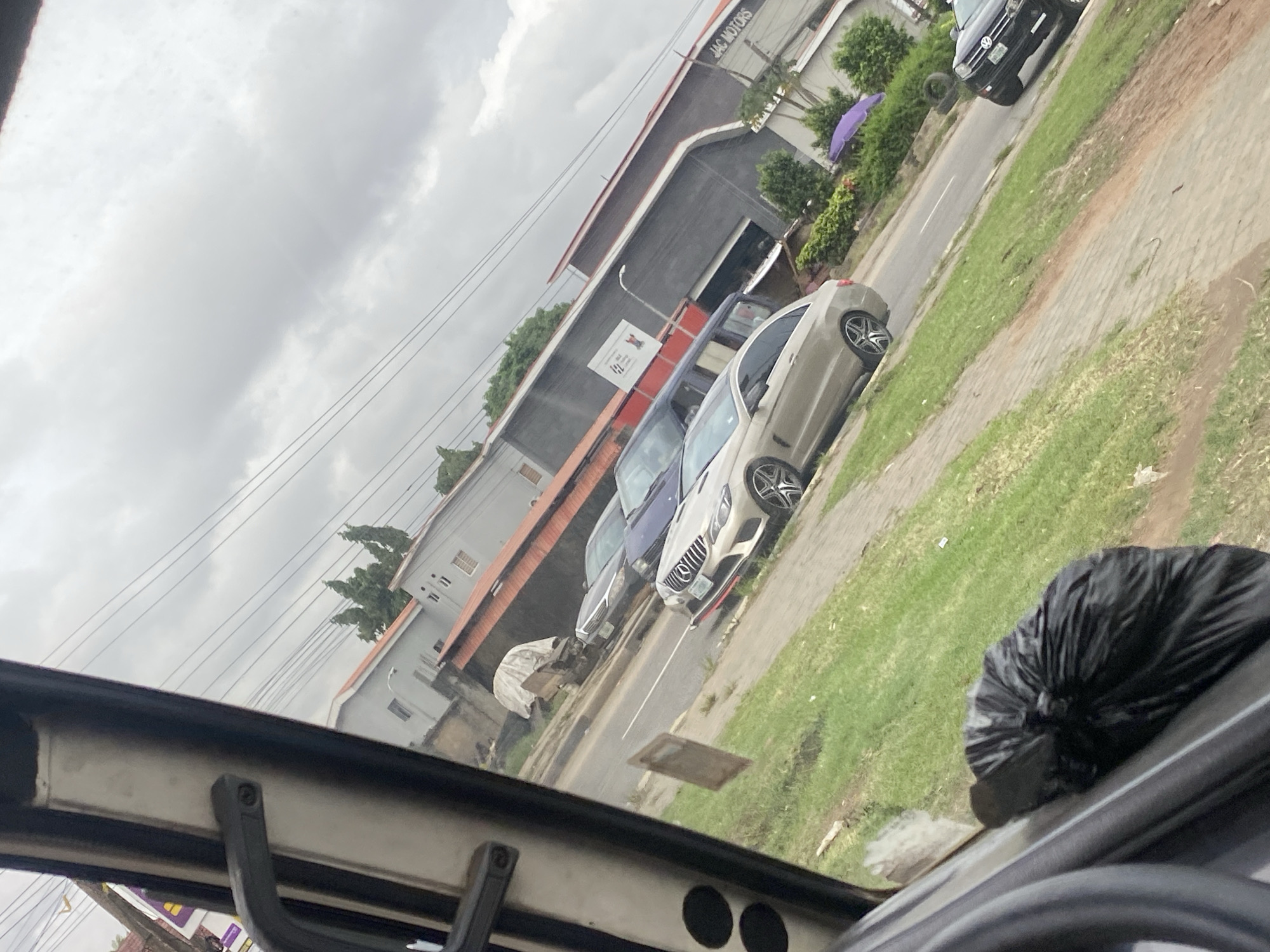 A side view of a street in Ikeja, Nigeria, shows a beige Mercedes-Benz E-Class coupe parked on the right side of the road. The car has a red trim below the front bumper and custom wheels. To the left of the Mercedes, a darker sedan is partially visible. In the background, a gray building with a red roof is seen, and further back, a longer gray building with "JAC MOTORS" written on its side. The sky is overcast. The foreground includes a black trash bag on the left side of the frame.