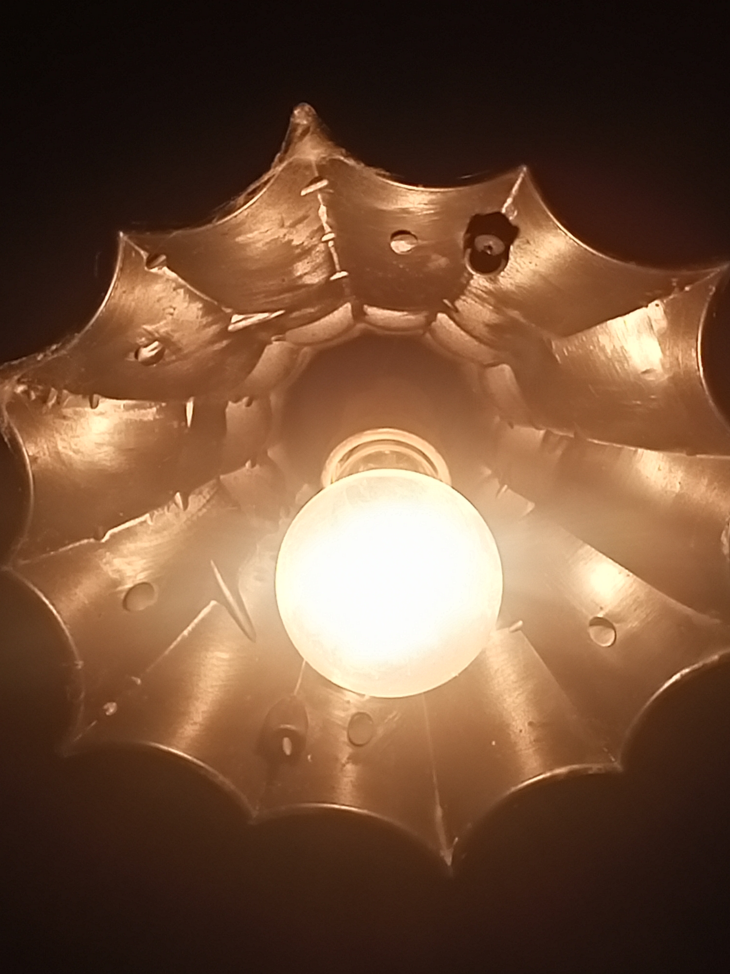 This is a close-up, indoor shot of a brass lamp fixture with a light bulb. The fixture has a multi-pointed, petal-like design, with visible seams and rivets holding the sections together. The surface of the fixture is textured and reflective, catching the light from the bulb. The bulb itself is illuminated and spherical, positioned centrally within the fixture. The background is dark and out of focus. There are no people or animals visible in the image. This is not an image of Sokuluk, Kyrgyzstan.