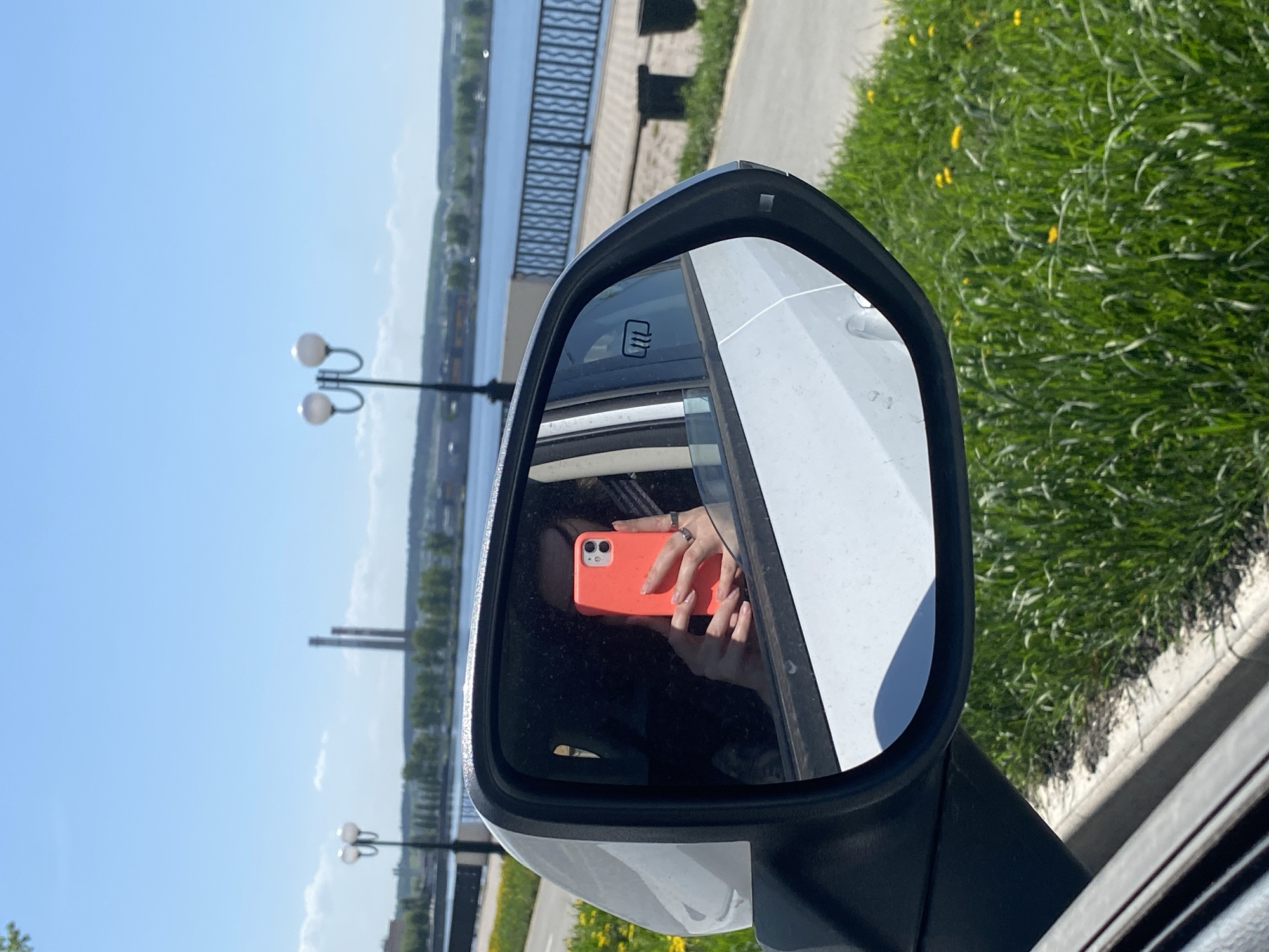 The image shows a car's side mirror reflecting a person taking a selfie with an orange smartphone. The mirror's background reveals a cityscape, a body of water (likely the Votkinsk Reservoir), streetlights, and green vegetation. The location is likely in Votkinsk, Russia. The car is parked near a curb, with grass growing along the edge.