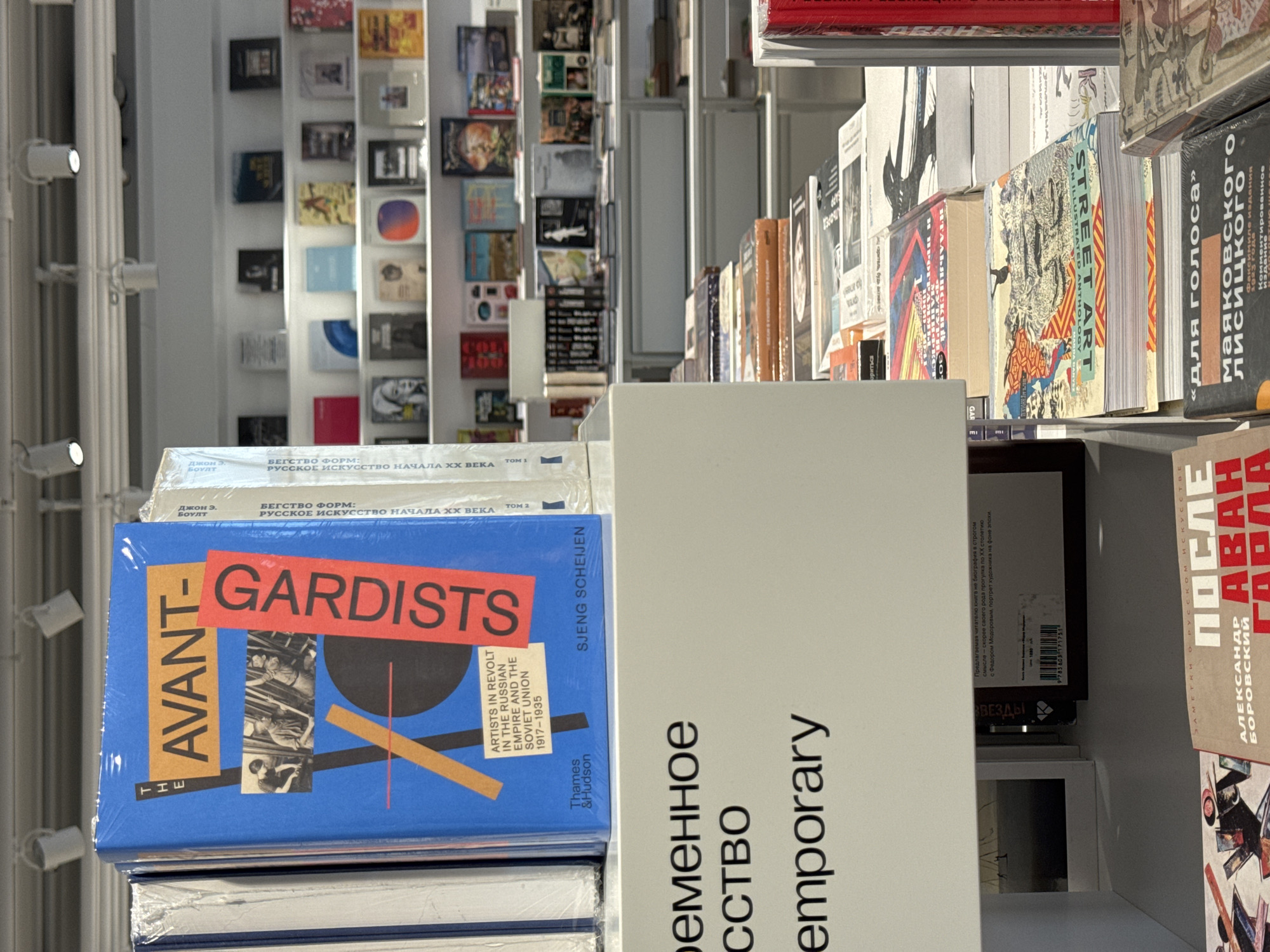 This is a close-up, indoor shot of a bookstore shelf, likely in Moscow, Russia, given the presence of Russian text on many of the books. The shelves are filled with books, a mix of both Russian and English titles, many related to art and design. A blue book titled "The Avant-Gardists" is prominent in the foreground, its cover featuring geometric shapes and text. To the right, a book with partially obscured Russian text is visible. The background shows additional shelves filled with books. The lighting is bright, illuminating the details of the book covers and spines.