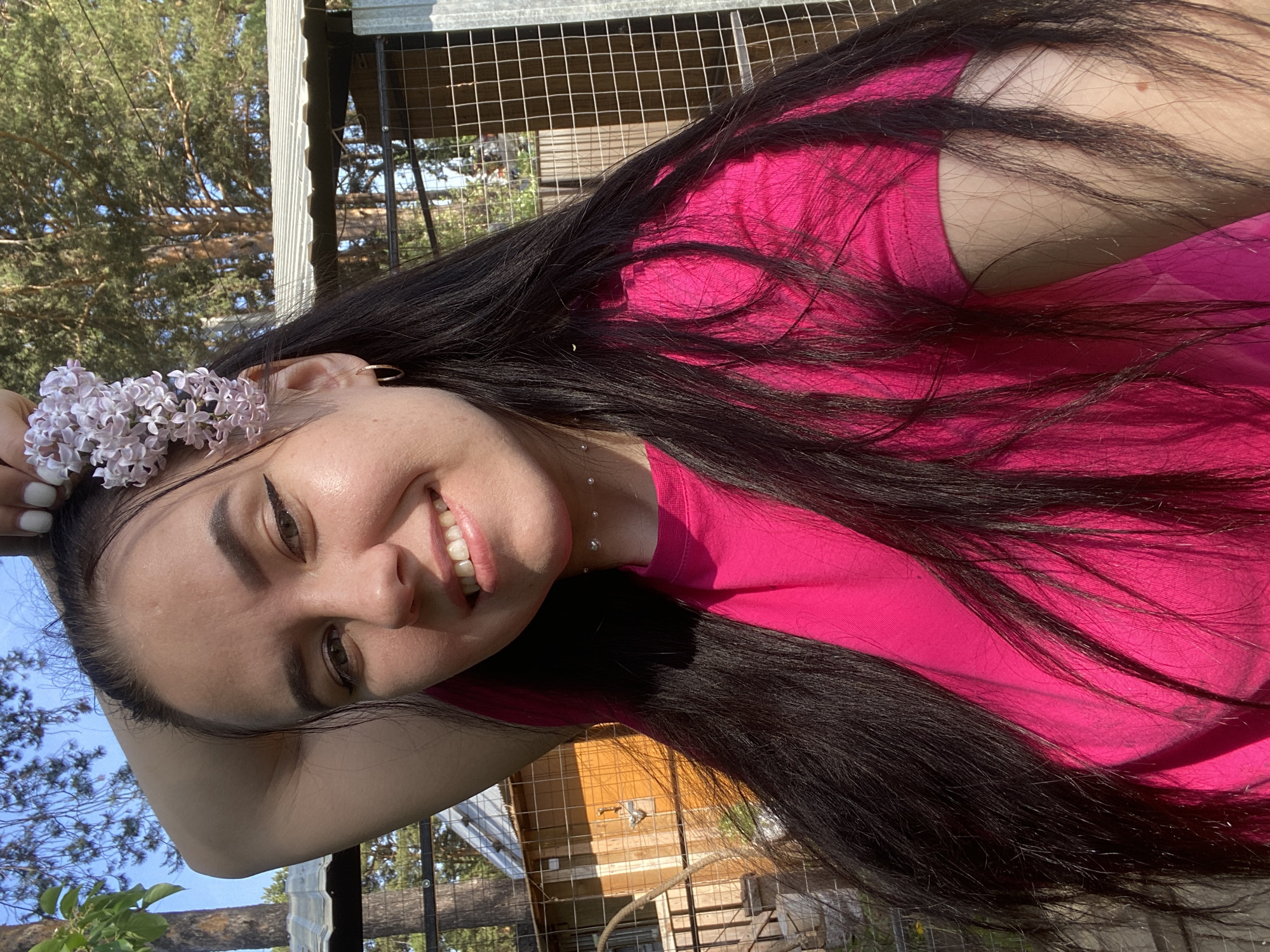 A woman with long, dark hair is smiling at the camera. She is wearing a bright pink t-shirt and has white flowers in her hair. She appears to be outdoors, as trees and a fence are visible in the background. This is in Chaikovsky, Russia.