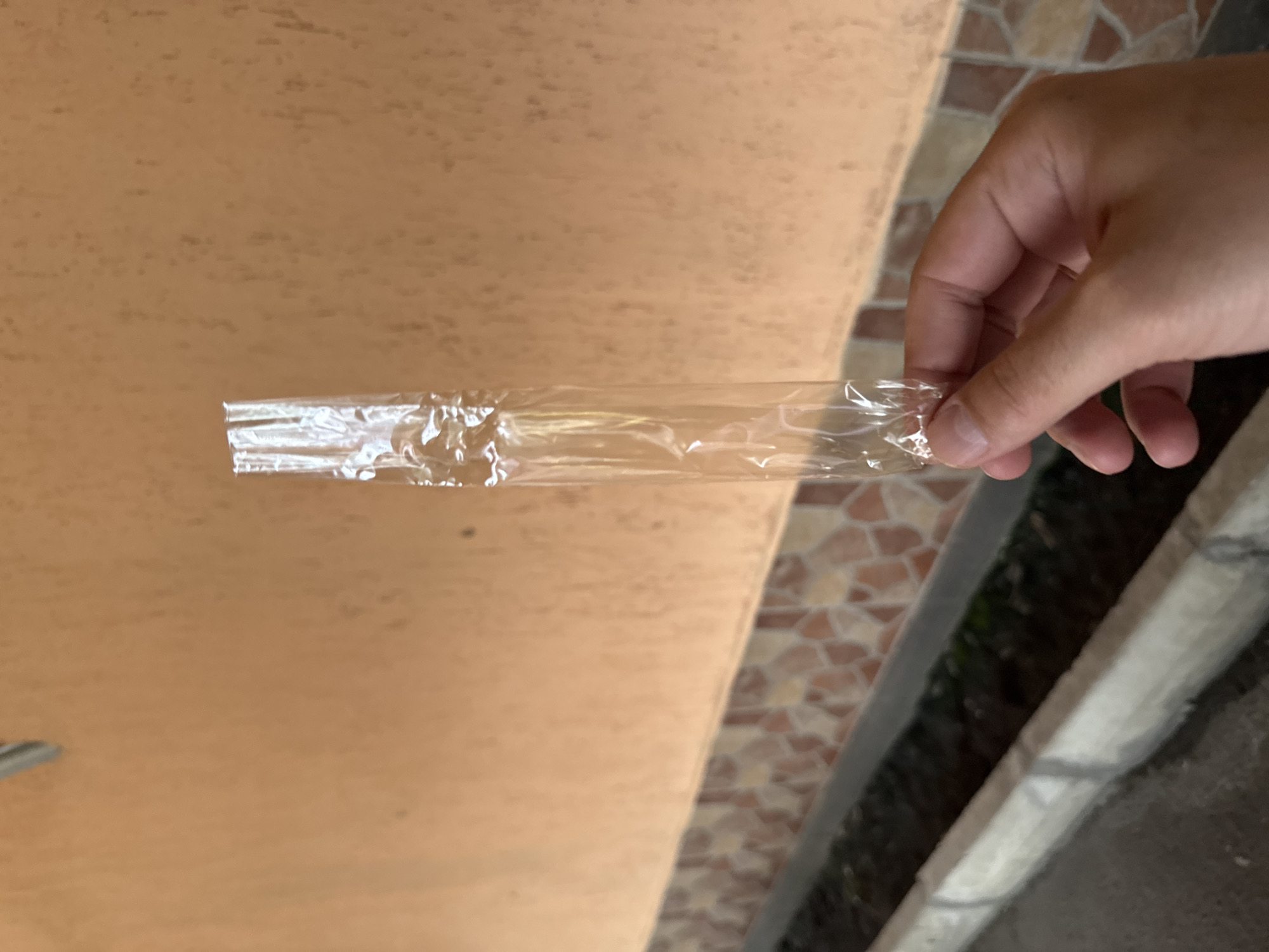A right human hand, light-skinned, is positioned in the lower-right foreground, holding a transparent, elongated plastic wrapper vertically between the thumb and index finger. The wrapper appears empty and crinkled, particularly at its upper end, suggesting it has been opened or sealed.
The background consists of an exterior wall structure. The upper portion of the wall is painted a solid peach or light orange color, featuring a subtly textured finish with scattered darker speckles. Below this, a horizontal band of decorative tiles covers the lower section of the wall. These tiles are irregularly shaped and feature a mosaic-like pattern in shades of reddish-brown, beige, and cream. Below the tiled section, a grey concrete curb or raised edge is visible, running horizontally across the lower part of the image. Beyond the curb, a narrow, dark, recessed area, possibly a planter bed or drainage channel, contains dark soil and faint glimpses of green vegetation. The lowest part of the scene shows a dark grey paved surface, likely a sidewalk or street.
The overall scene depicts an outdoor urban environment. While the prompt specifies the location as Sofia, Bulgaria, there are no discernible unique visual cues within the image itself (such as specific landmarks, street signs, or highly distinctive architectural features) to forensically pinpoint a precise location within the city of Sofia.