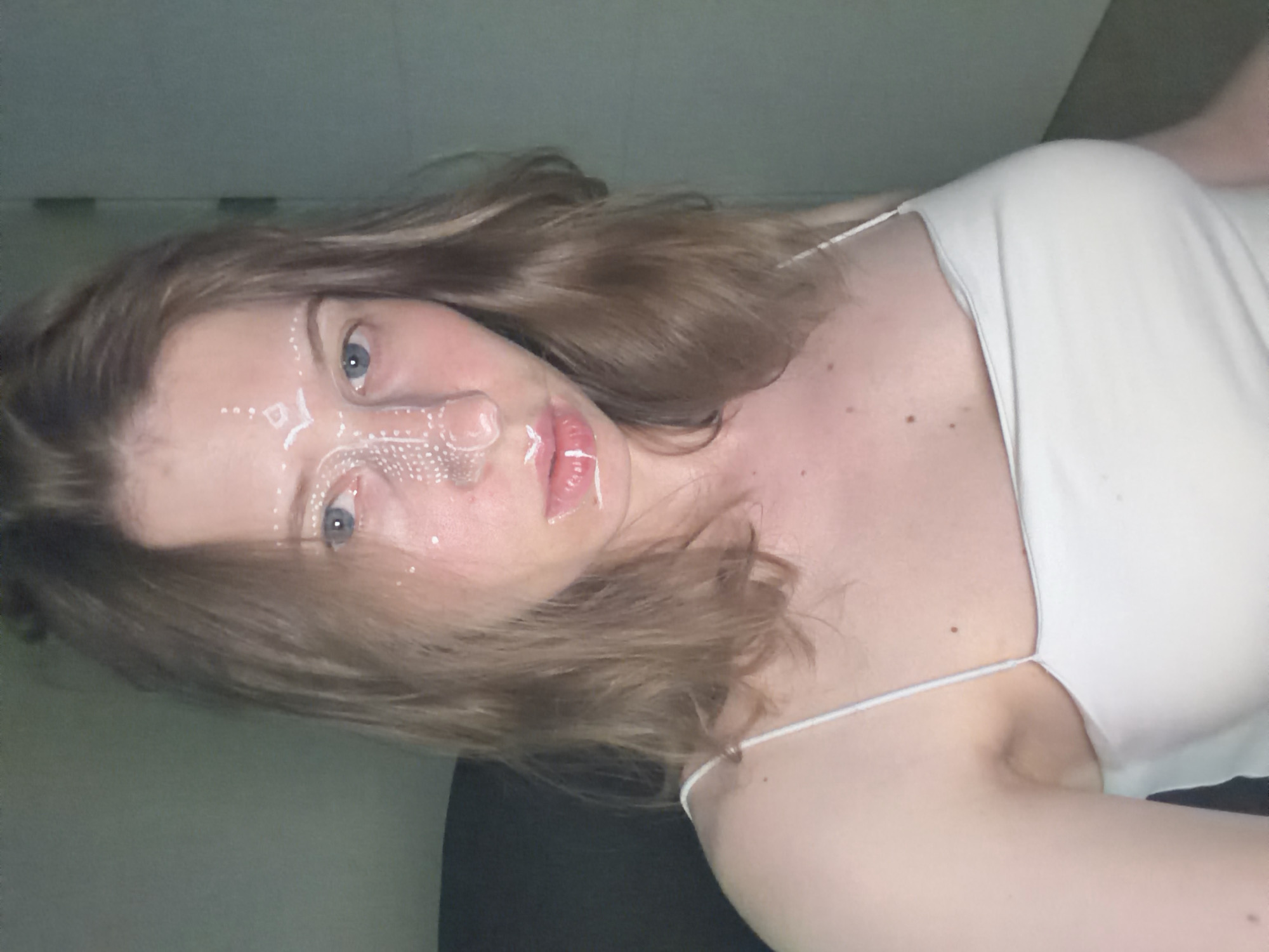 A young adult with light brown hair and blue eyes, possibly female, is pictured in a dimly lit room. They are wearing a white tank top and have intricate white face paint or makeup applied to their forehead, nose, and lips. The background appears to be a wall, with a dark object, possibly a chair, visible on the left. There is no discernible location in Dongducheon-si, South Korea.