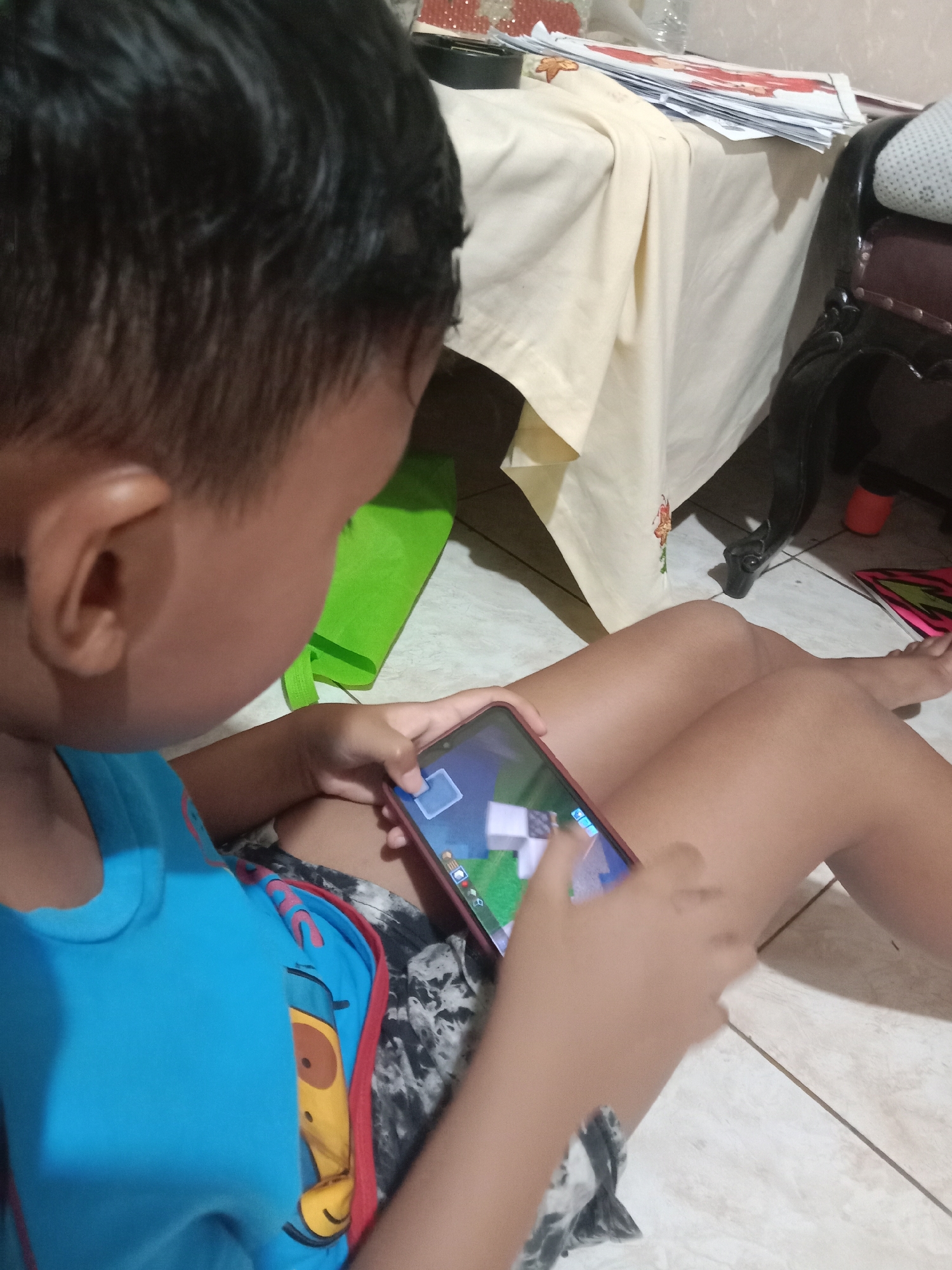 A young child, seen from the back, is playing a video game on a smartphone. The child is wearing a blue t-shirt and patterned shorts, and is sitting on a tiled floor. Their legs are crossed, and their hands are occupied with the phone. In the background, a beige tablecloth covers a table, with some papers on top. A green bag is visible behind the child. The overall lighting suggests an indoor setting, likely a home environment in Madiun, Indonesia.