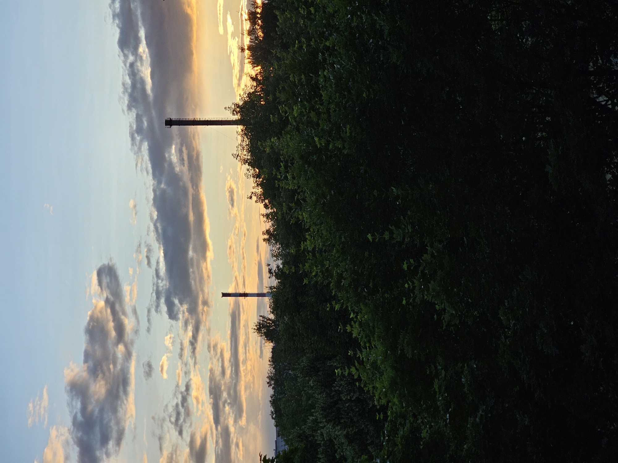 The image depicts a dusk scene in the city of Khimki, Russia, characterized by a silhouetted line of lush green trees in the foreground. Behind the trees, two industrial chimneys stand tall against a sky illuminated by a warm sunset glow. The sky is a gradient of light blue transitioning to shades of orange and yellow near the horizon, with scattered clouds tinged by the sunset. The overall impression is one of a serene, possibly industrial, landscape at the end of the day.
