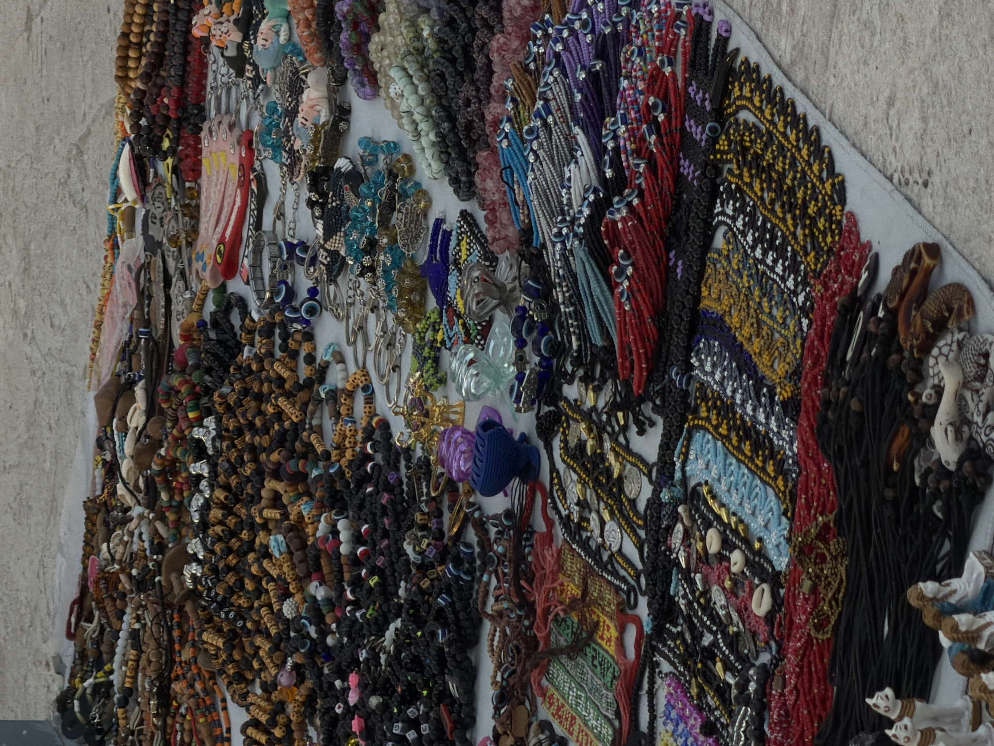 The image displays a vibrant and diverse collection of handmade jewelry, likely a street vendor's stall in Sharm El Sheikh, Egypt. The jewelry is spread out on a light-colored surface, possibly a rug or cloth, on top of a textured, light-colored ground. The majority of the items are necklaces and bracelets, featuring a wide array of beads in various materials and colors, including wood, glass, and stone. Many pieces incorporate intricate patterns and designs, such as geometric shapes and stylized motifs. Several pieces prominently feature blue and red beads, with some also including "evil eye" charms, a common protective talisman. Among the jewelry, small figurines of cats and human-like figures are also visible, adding to the eclectic nature of the display. The overall impression is one of abundance and variety, characteristic of a bustling market or tourist area.