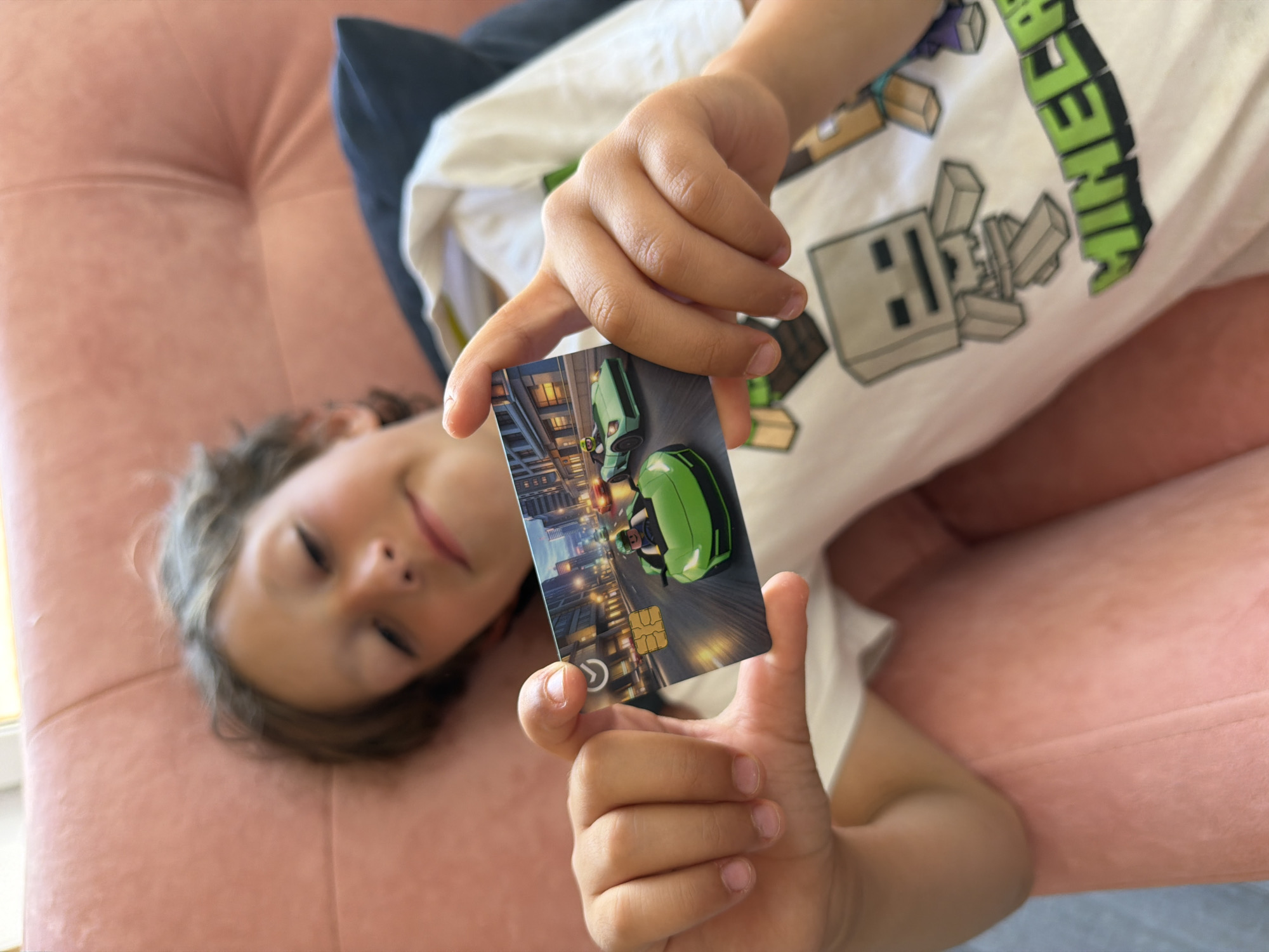 A young child lies on a pink tufted sofa, looking up at the camera with their eyes closed and a slight smile on their face. The child is wearing a white t-shirt with a Minecraft graphic. Their right hand is raised, holding a card with an image of a neon green futuristic car in a city setting. The card has a gold chip and a circular logo. The setting appears to be indoors, possibly a living room, with natural light suggesting daytime. The overall mood is relaxed and playful.