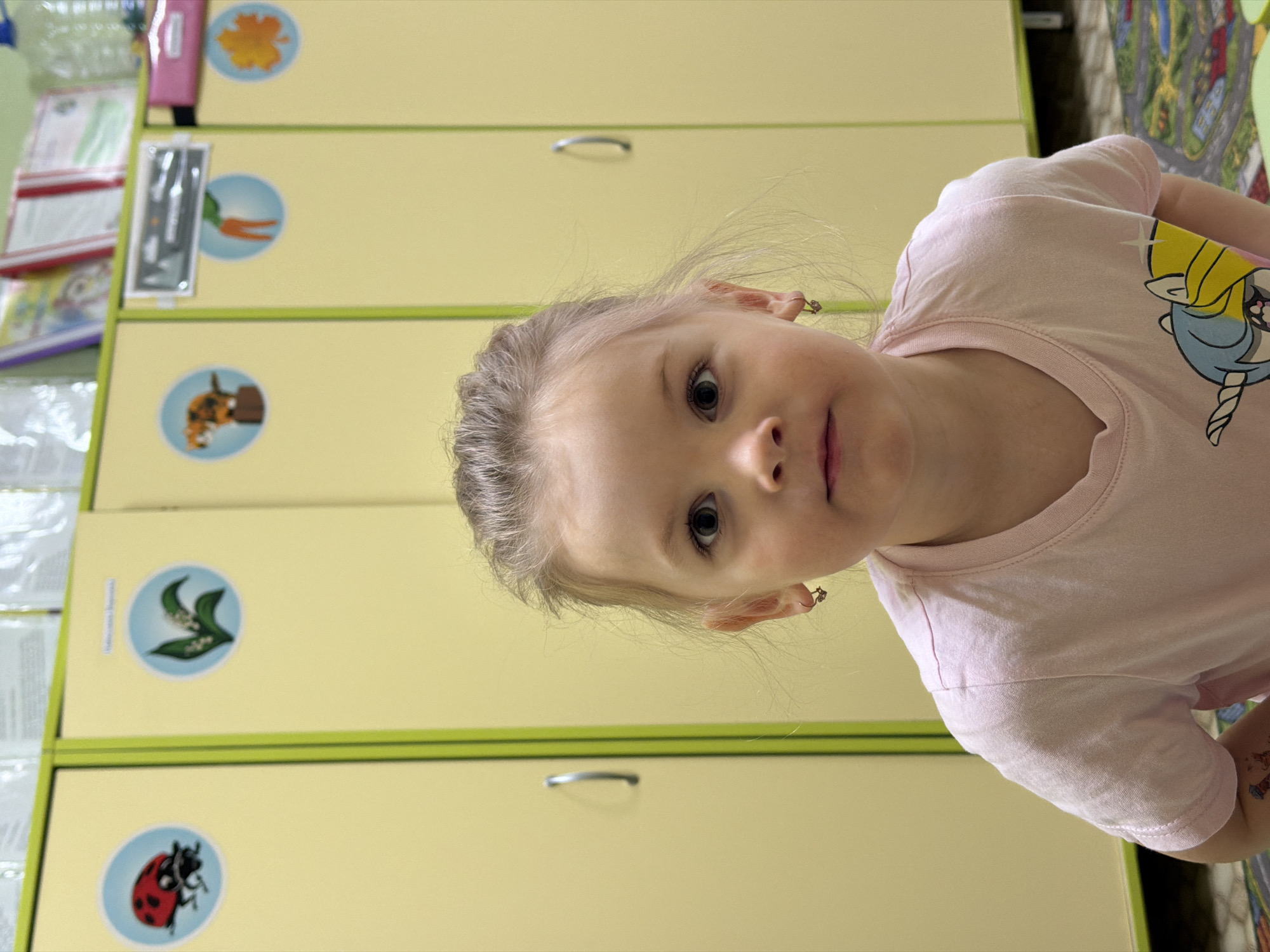 The image, rotated 90 degrees clockwise, features a young girl in an indoor setting, likely a playroom or classroom in Chaikovsky, Russia. She is depicted in a close-up portrait, looking directly at the camera with wide, attentive eyes and a neutral to slightly curious expression. The girl has light-colored hair and is wearing a light pink short-sleeved t-shirt, which features a cartoon unicorn or pegasus design on the right chest/shoulder area. Small, delicate earrings are visible in her ears.
The background consists of tall, light yellow storage cabinets with bright green trim, characteristic of a child-focused environment. These cabinets are decorated with several circular decals, each illustrating a different item. From top to bottom, visible decals include an orange leaf or flower, a carrot, a squirrel next to a nut, lilies of the valley, and a ladybug. On the far left, shelves hold various items like papers or books. A colorful playmat, possibly depicting roads or a town, is partially visible on the floor to the right. The scene suggests a cheerful and organized space designed for children's activities.