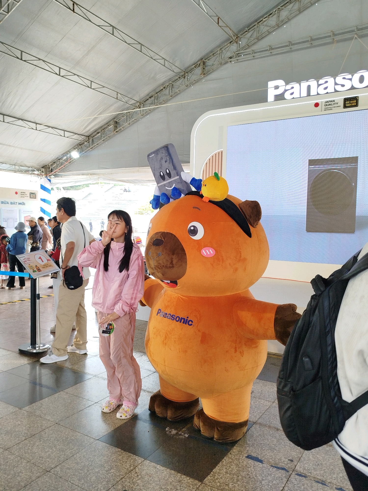 An outdoor event, possibly at a Panasonic exhibition, is captured on a sunny day under a large white tent structure. In the foreground, a young Vietnamese girl in pink athletic wear poses with a peace sign towards the camera. She stands beside a large, inflatable mascot resembling an orange bear with brown ears and paws, wearing a black eye patch and a small yellow fruit-like accessory on its head. The mascot has "Panasonic" written on its chest in blue. To the left of the girl, a man in a white polo shirt and beige pants stands with a black bag slung over his shoulder. Behind them, other attendees are visible, including children and adults. A display board with Vietnamese text and images is positioned to the left. The floor is paved with square tiles. In the background, a large screen displays a Panasonic logo and an image of a washing machine. The overall atmosphere suggests a promotional event in a public space, likely within Da Lat, Vietnam, given the Panasonic branding and the presence of local signage and people.