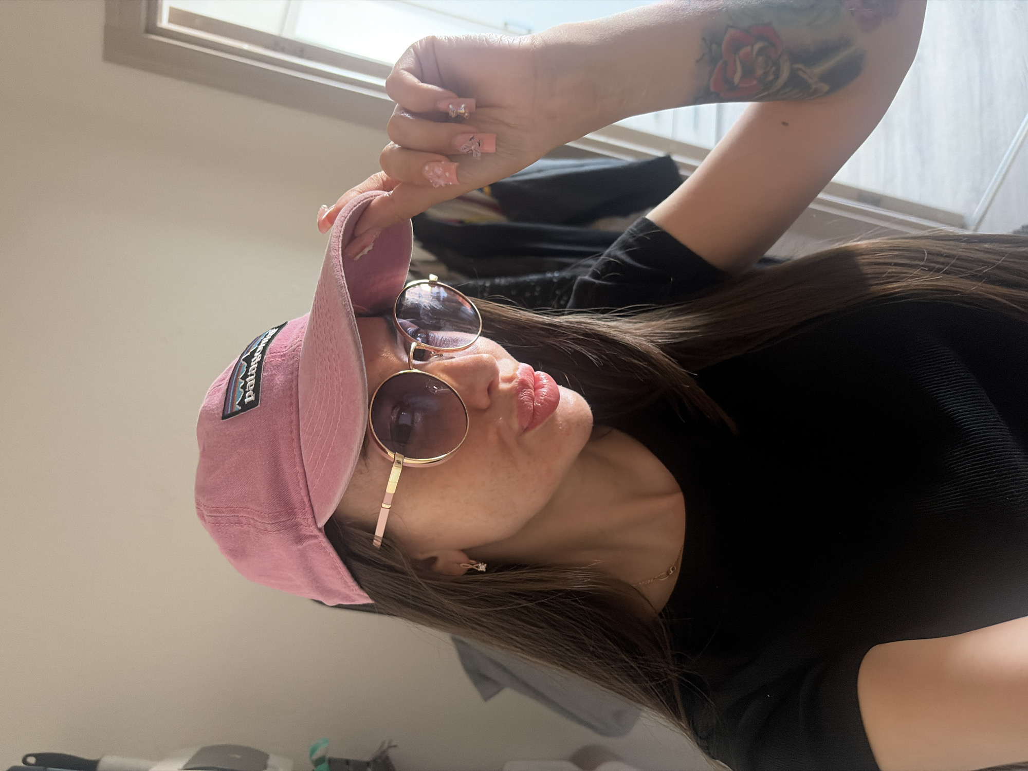 The media file contains a close-up, vertical, shot of a person wearing a pink baseball cap and round sunglasses. The person has long, dark brown hair and is wearing a black top. Their left arm is visible, and a tattoo of a red rose is on their upper arm. The person's nails are long and decorated with pink and white designs, including small floral embellishments.
The person is looking to their left and appears to be posing. Their lips are slightly parted, and they are wearing subtle makeup. A thin gold chain necklace is visible around their neck. The background is softly blurred, showing what appears to be an indoor setting with light-colored walls and a window with blinds. There is also a hint of dark clothing in the background, possibly hanging items.
A visible logo on the pink cap reads "PATAGONIA" within a colorful rectangular patch. The lighting suggests it is daytime. The overall mood appears casual and self-assured.