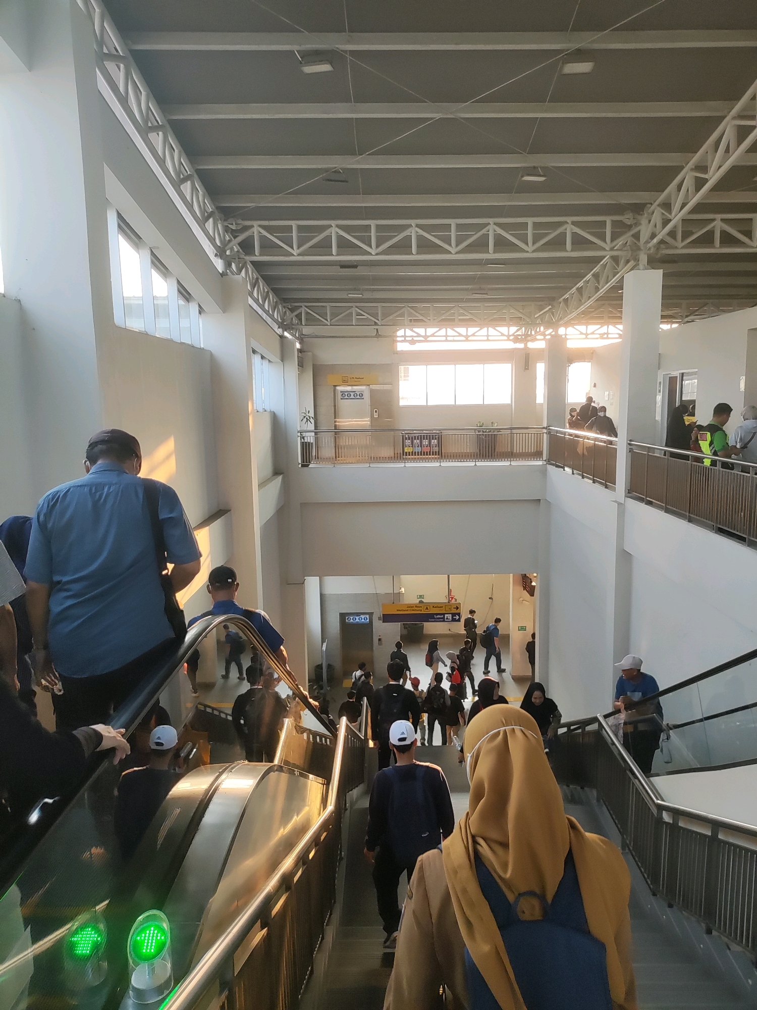 The image depicts the interior of a modern building, likely a transit station or mall, with a bright, open-air design featuring large windows and a high ceiling with exposed industrial trusses. The architecture suggests a contemporary urban environment, possibly in Telagamurni, Indonesia, given the presence of what appears to be signage with Indonesian text.
In the foreground, several people are moving up an escalator. The person closest to the camera, on the left side of the escalator, is wearing a blue short-sleeved shirt and a dark bag slung over their shoulder, moving upwards. To their right, another individual in a blue shirt and dark cap is also ascending. Further down the escalator, more people are visible, some with backpacks and casual attire. The escalator itself is metallic with a dark railing, and a green arrow indicating the direction of travel is illuminated.
On the left side of the image, a white wall with large windows lets in ample natural light, casting warm shadows. On the right side, another escalator is visible, with people descending. In the mid-ground, there's a lower level with more people walking and congregating. Signs are present, including one with the text "Lift Kanan" which translates to "Right Lift". Another sign in yellow with black text and arrows indicates directions, with the phrase "Jalan Umum" (General Street) and "Masjid" (Mosque) potentially visible.
The overall atmosphere is one of movement and activity, with individuals engaging in travel or transit. The environment is clean and well-lit, with a clear focus on public accessibility and flow.