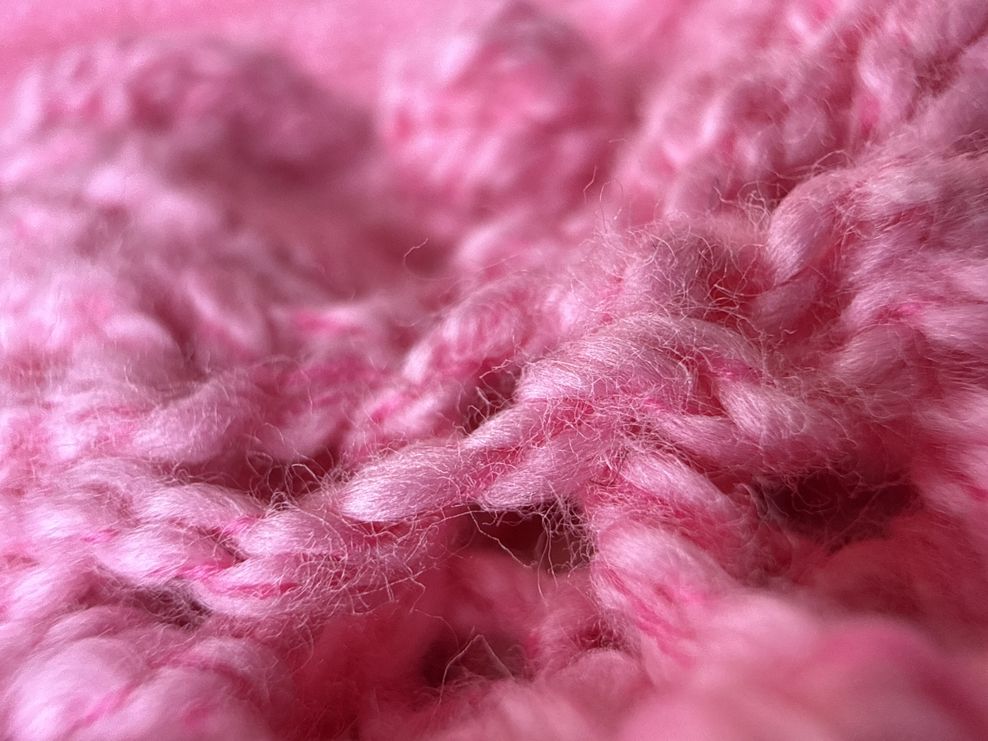The image is a close-up, macro photograph of a textured pink knitted fabric. The focus is sharp on the intricate weave of the yarn, revealing individual fibers and the raised loops of the knit. There are variations in the shade of pink, with some areas appearing brighter and more saturated than others, suggesting either a multi-toned yarn or subtle lighting differences. The texture is soft and fuzzy, with some loose fibers protruding from the main structure of the knit. The background is blurred, but also appears to be the same or a similar pink knitted material, creating a monochromatic and soft aesthetic. There are no people, discernible objects, or environmental features visible in the image. The image does not provide any information that would allow for its location to be identified in Moscow, Russia or anywhere else.
