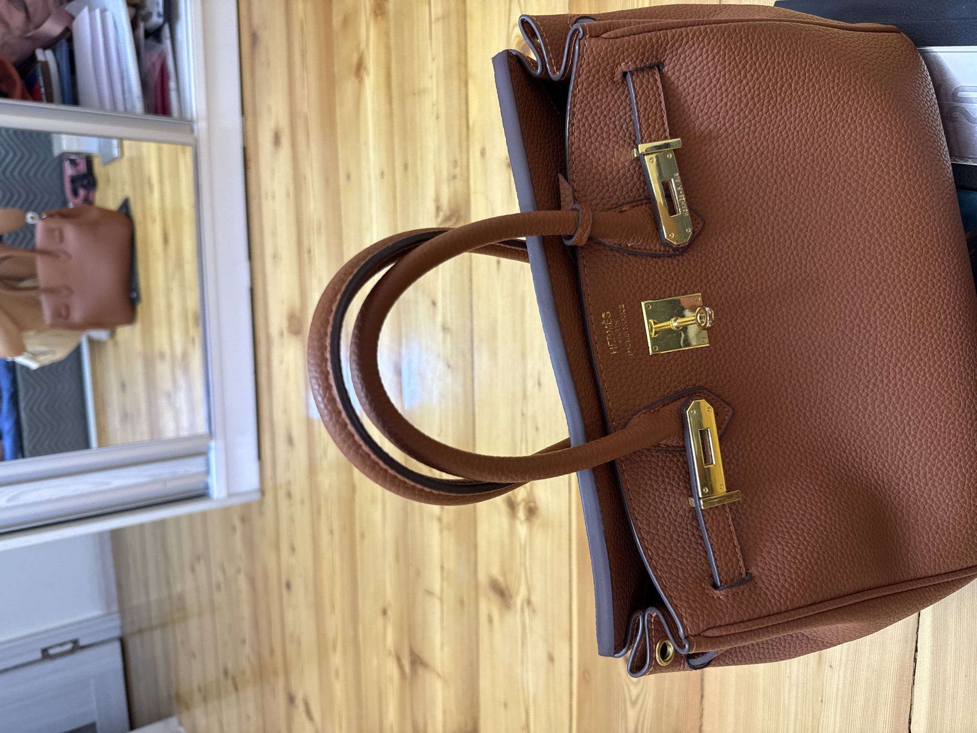 A brown pebbled leather handbag with gold hardware is centered in the foreground, positioned on a light brown wooden floor. The handbag features two top handles, a flap closure with a turn-lock mechanism, and prominent stitching. Embossed text on the flap reads "HERMÈS PARIS MADE IN FRANCE". In the background, a large mirror reflects a blurry scene of a room, including a person holding a similar handbag and a dresser with stacked items. The environment appears to be indoors, possibly a bedroom or dressing area, with a wardrobe or closet visible to the right. The lighting is soft and diffused, suggesting natural light or indoor ambient lighting. The image quality is high, with a shallow depth of field that blurs the background. There are no visible signs or indications that this image was taken in Tashkent, Uzbekistan.