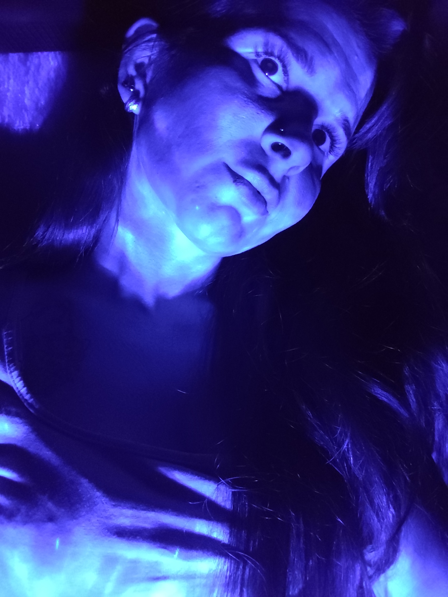 A young woman is illuminated by a strong blue light in an indoor setting. She has long, dark hair and is wearing a casual top. Her face is tilted slightly, and her eyes are wide. A small nose piercing is visible. The background is dark and indistinct. There is no information to suggest that the image was taken in Фоки, Russia.
