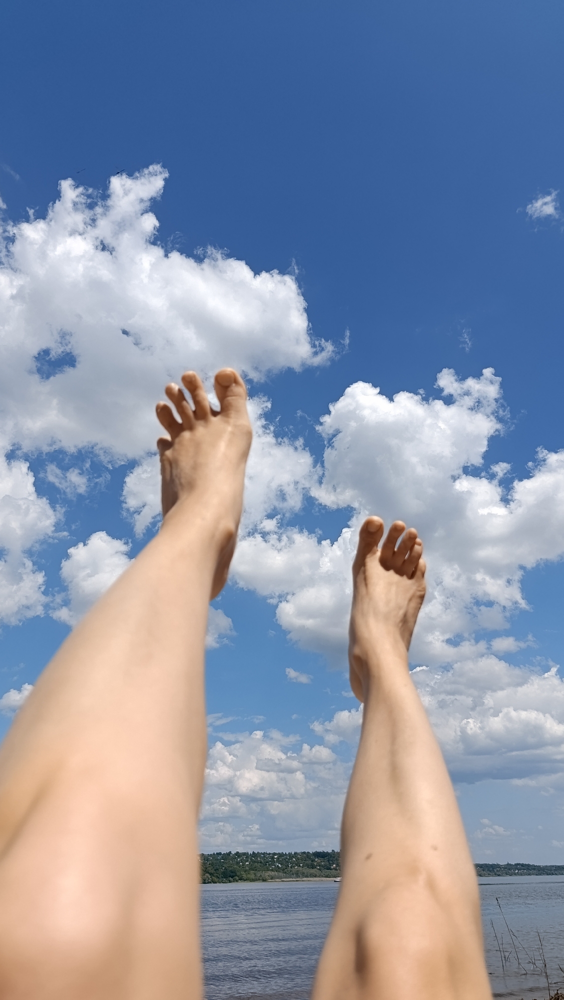 The image shows a pair of bare feet and lower legs held up against a bright blue sky filled with white, puffy clouds. In the background, a body of water, likely a river, is visible, with a green, tree-lined shore and a distant settlement. The perspective is from below, looking up at the legs and sky. Without additional information or visual cues specifically pointing to Saratov, Russia, it is impossible to confirm the location. The scene appears to be outdoors on a sunny day, suggesting a recreational setting. There are no people or objects clearly identifiable in the frame other than the legs and feet, the sky, clouds, and the distant landscape.
