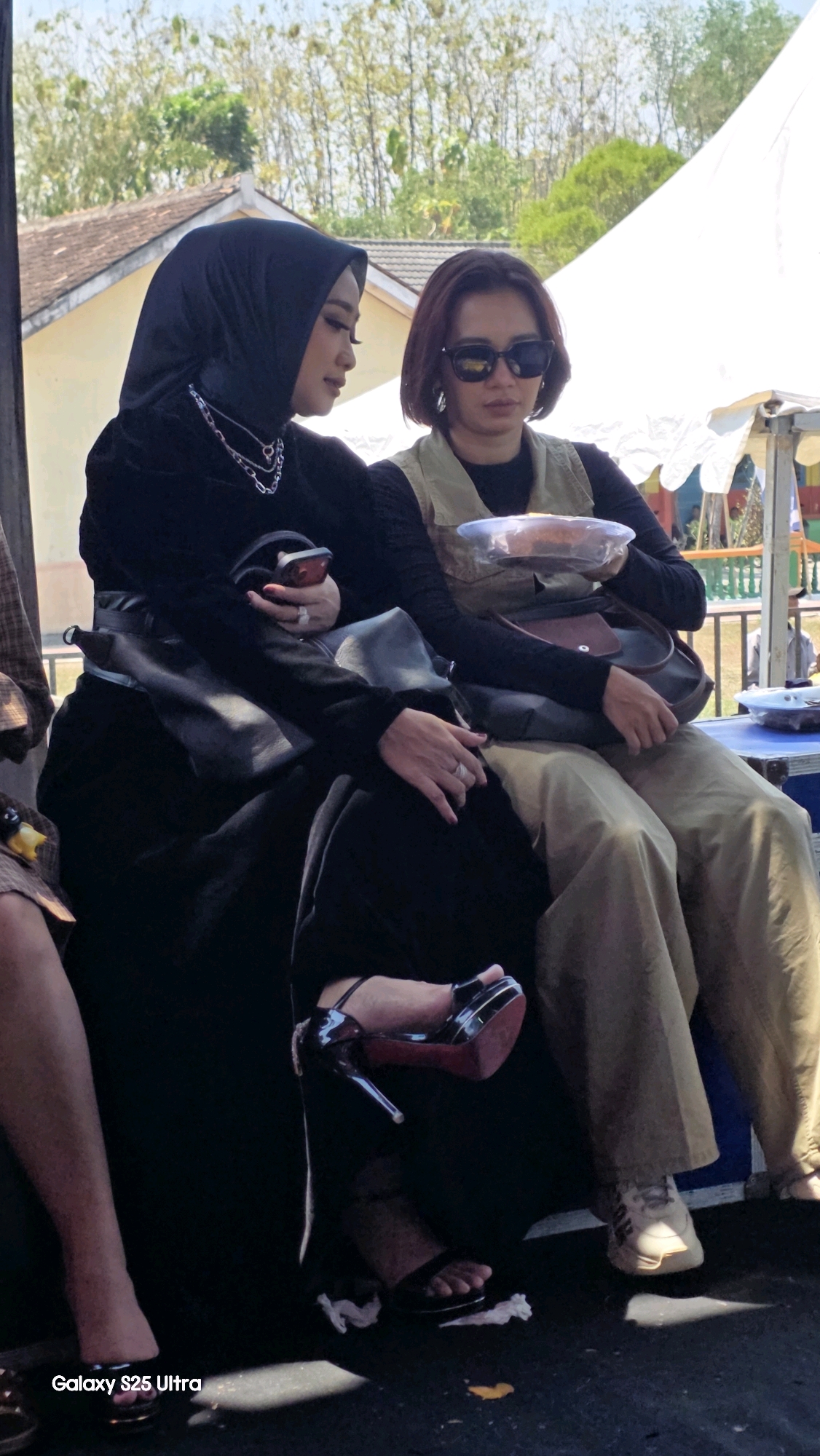 Two women are seated outdoors, appearing to be at a community event or market in Toroh, Indonesia. The woman on the left wears a black hijab and a black outfit, accessorized with a silver chain necklace and rings. She holds a smartphone in her hand and a dark handbag rests on her lap, partially obscuring her black dress. Her feet are adorned with black high-heeled sandals. The woman on the right has short, dark hair and wears sunglasses. She is dressed in a beige vest over a black top and loose-fitting tan pants. A handbag is slung across her body, and she holds a clear plastic container with food. Her feet are clad in white sneakers. Behind them, a white tent is visible, along with the roof of a building and trees in the background, suggesting an outdoor setting under a bright sky.