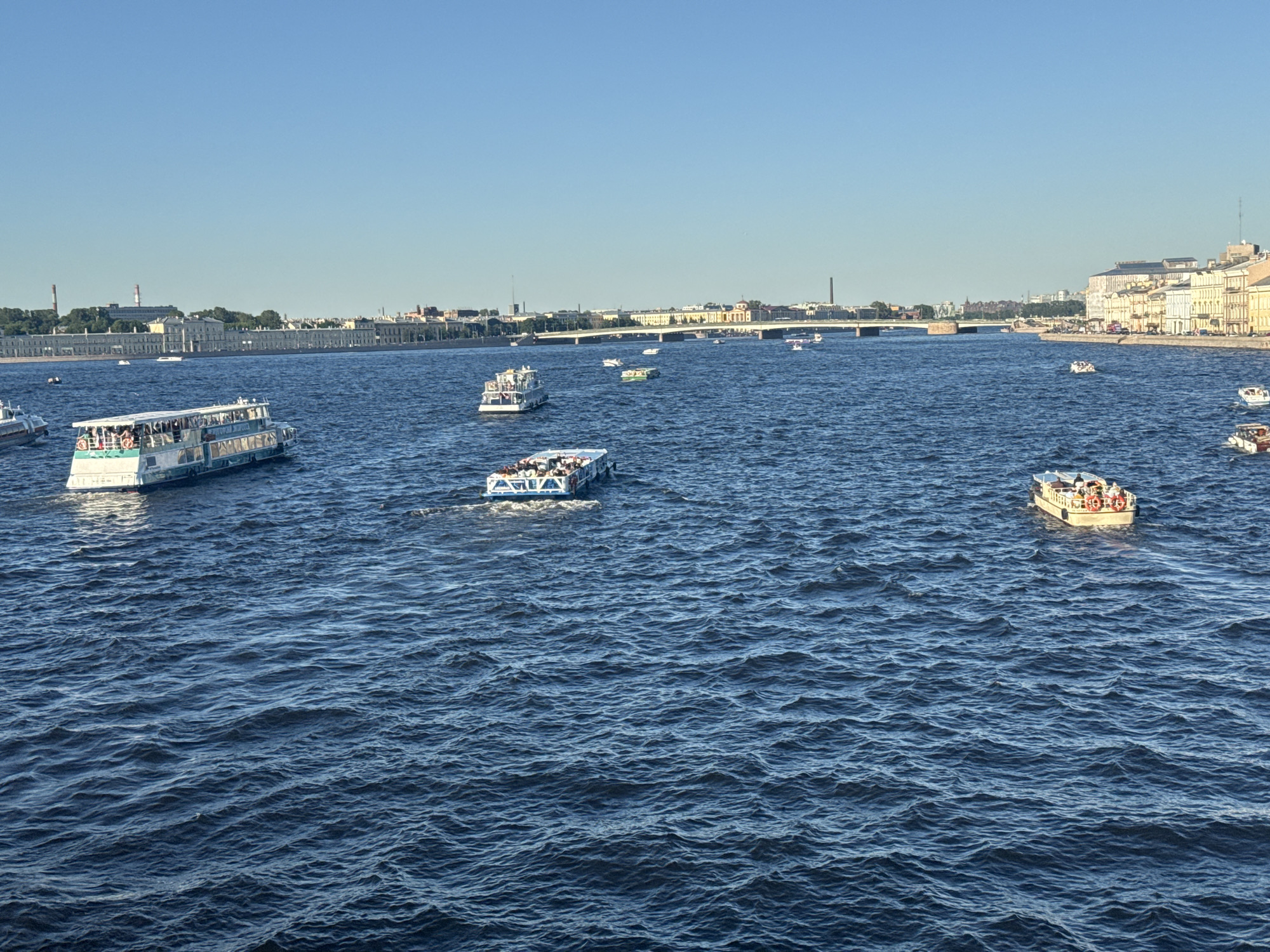 The image depicts a wide river in Saint Petersburg, Russia, with several tour boats navigating its waters. The sky is clear and blue, suggesting a sunny day. The riverbanks are lined with historical buildings and a bridge spans the waterway in the distance. The boats appear to be carrying passengers, likely tourists enjoying a scenic tour of the city. The water is rippled, indicating a gentle breeze. The overall atmosphere is one of a pleasant summer day with typical city life activity on the river.